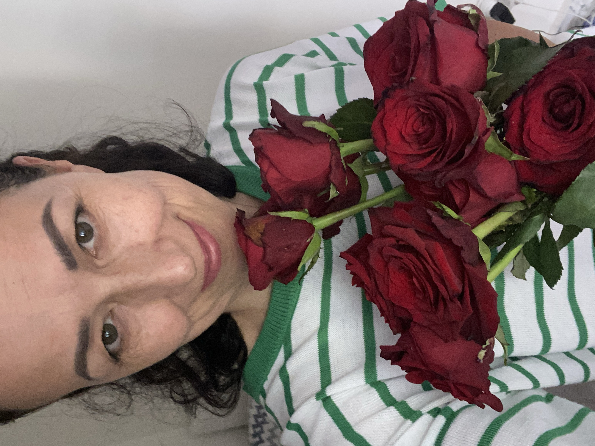 A middle-aged woman with dark hair, light skin, and brown eyes smiles while holding a bouquet of dark red roses. She is wearing a white and green striped shirt. The photo appears to be taken indoors. There is no information in the image to suggest that the location is Alanya, Turkey.