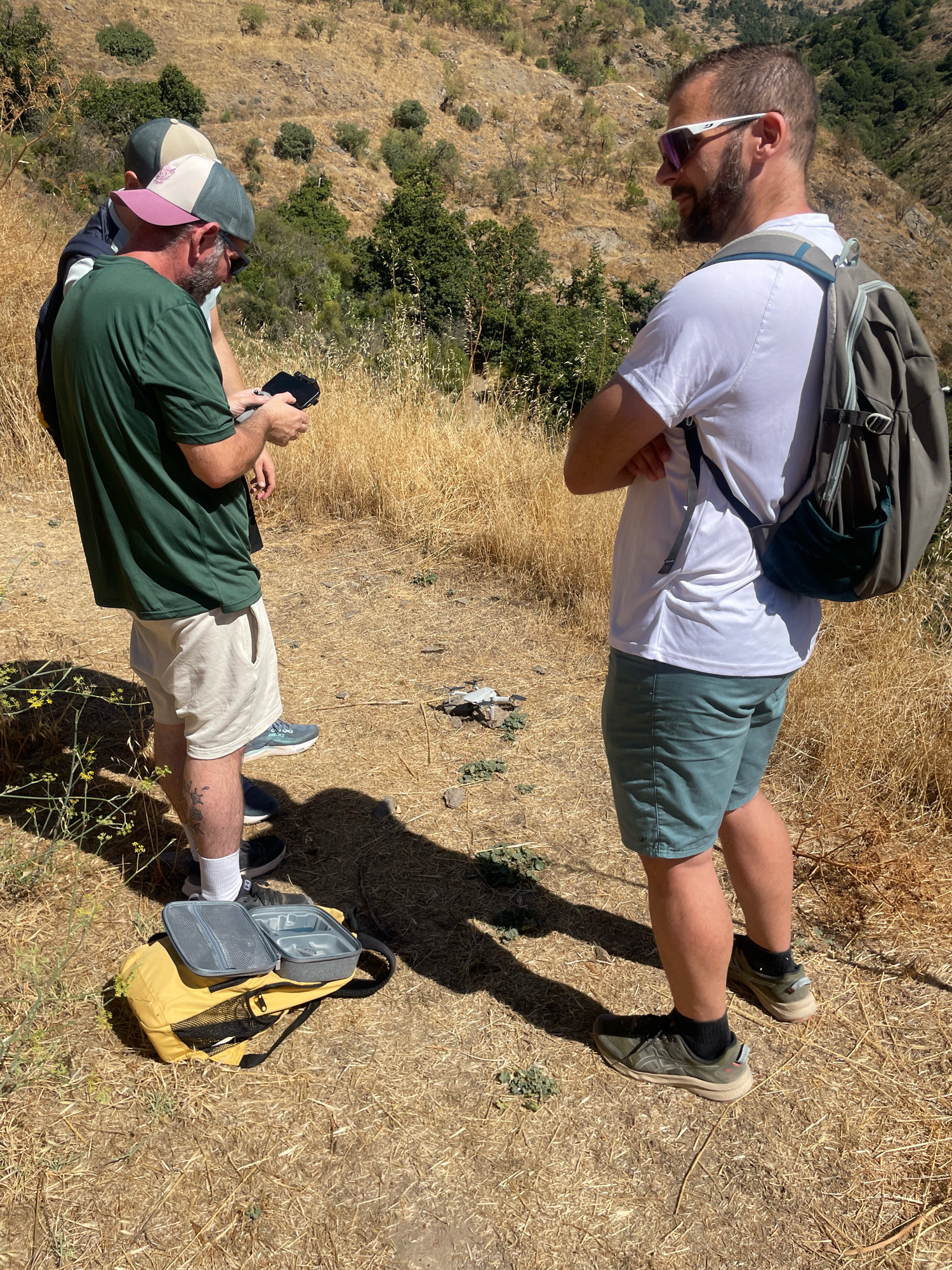 Three men stand in a dry, grassy, mountainous area. Two men are in the foreground on the left, one wearing a green t-shirt and light-colored shorts, and the other partially visible wearing a dark shirt and a baseball cap. The man in the green t-shirt is holding a black object, possibly a phone, and is looking down at it. A yellow backpack with an open grey lunchbox is on the ground in front of them. A third man is on the right side of the image, facing away from the camera, wearing a white t-shirt, teal shorts, and a backpack. He has on sunglasses and appears to be looking to the left. The environment is arid with dry grass, scattered shrubs, and a steep, rocky hillside in the background. The lighting suggests it is daytime, likely sunny. The scene appears to be in an outdoor, natural setting, possibly a hiking trail in or near Lanjarón, Spain, given the dry, mountainous terrain characteristic of the region.