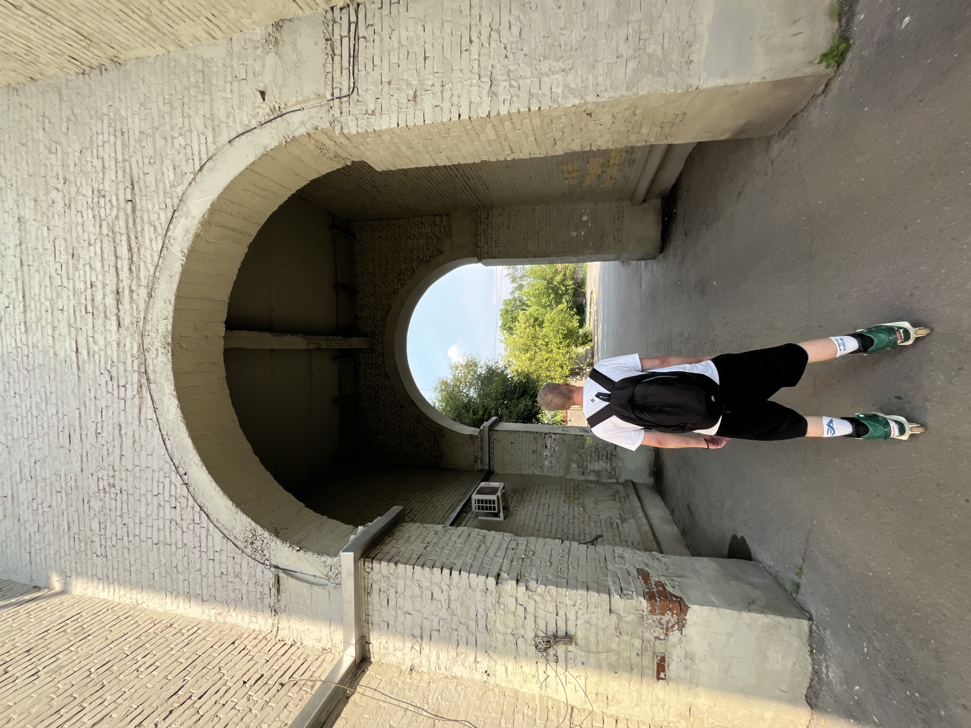 The image depicts a young man from the back, wearing a white t-shirt, black shorts, a black backpack, and rollerblades. He is standing in an arched passageway made of aged, painted brick. Sunlight casts a shadow on the left side of the passageway, highlighting the texture of the bricks. An air conditioning unit is mounted on the wall inside the archway. Beyond the arch, trees and a clear sky are visible. The scene appears to be an outdoor urban environment. It is not possible to determine the specific location in Krasnoyarsk, Russia, from the image alone.