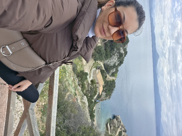 The image depicts a female subject in a selfie-style composition from an elevated viewpoint in the Municipal Unit of Loutraki - Perachora, Greece. She is positioned in the left foreground, wearing dark brown-tinted sunglasses, a light blue turtleneck, and a brown quilted puffer jacket. A beige shoulder bag with a silver D-ring buckle is draped across her chest. Her dark hair is pulled back.
Below her, a rocky, vegetated hillside descends steeply towards a large body of water. A rustic wooden railing is visible in the lower right foreground. In the midground, a small, light-colored building with a terracotta roof stands on a promontory, accessible by a winding dirt path. To the right of the building, a secluded cove with clear, light blue water and a small beach is visible, bordered by rocky cliffs; a small Greek flag is mounted near the building. The background features a vast expanse of calm, deep blue water leading to a distant mountainous coastline under a predominantly cloudy, grey sky.