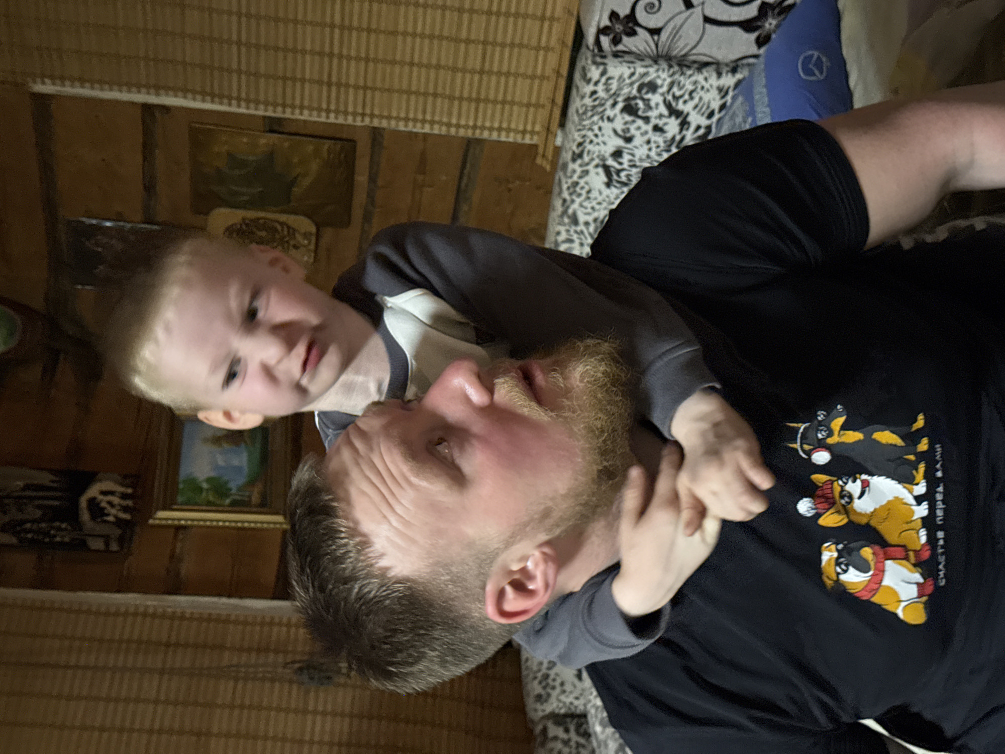 A man and a young child are indoors, interacting closely. The man, who appears to be lying on his back on a patterned surface (possibly a couch), has a light beard and is wearing a black t-shirt with a graphic featuring three stylized dogs. The child is on top of the man, facing the viewer with a somewhat mischievous expression. The child is wearing a two-tone grey and white long-sleeved shirt.
The background shows a room with wooden walls and patterned blinds. There are framed pictures or artwork on the wall. A patterned pillow is also visible on the surface the man is lying on.
There is no verifiable information to state this happened in Rosstani, Russia.