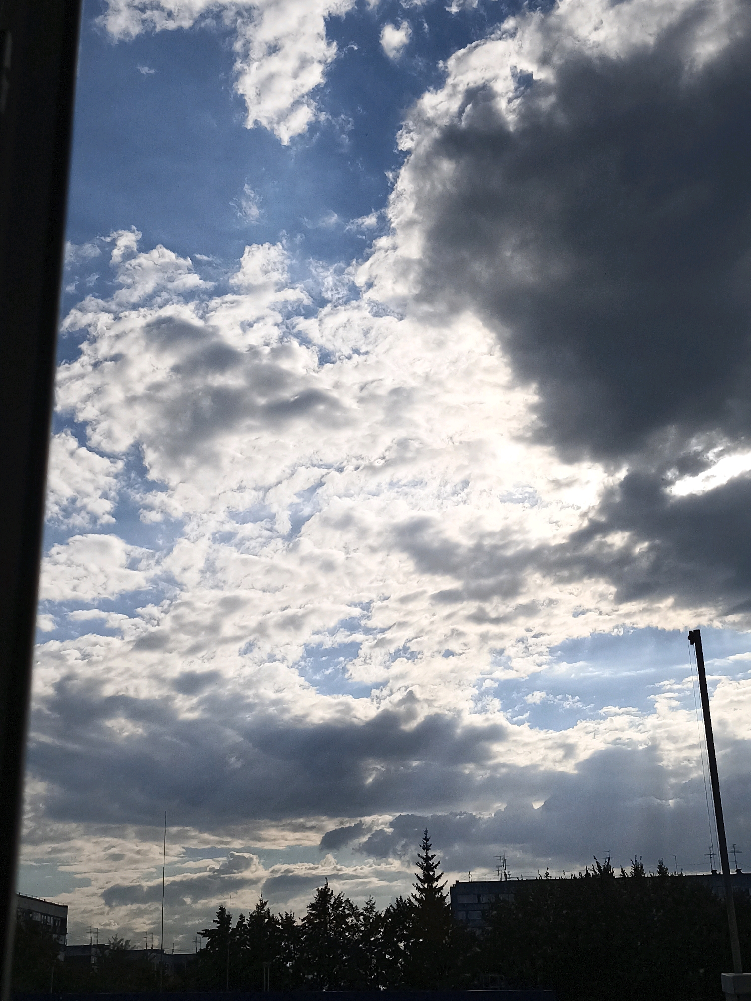 The image depicts a cloudy sky over a silhouetted cityscape. Dark, heavy clouds dominate the upper right portion of the frame, with patches of lighter, broken clouds interspersed with blue sky elsewhere. Sunlight filters through the clouds, creating bright areas. Below the sky, the silhouettes of trees, buildings, and a tall pole with a wire are visible against the brighter sky. The scene appears to be captured from an elevated viewpoint, possibly a window, given the dark, vertical frame on the left edge of the image. The time of day is likely late afternoon or early evening, indicated by the dramatic cloud formations and the way light is breaking through.