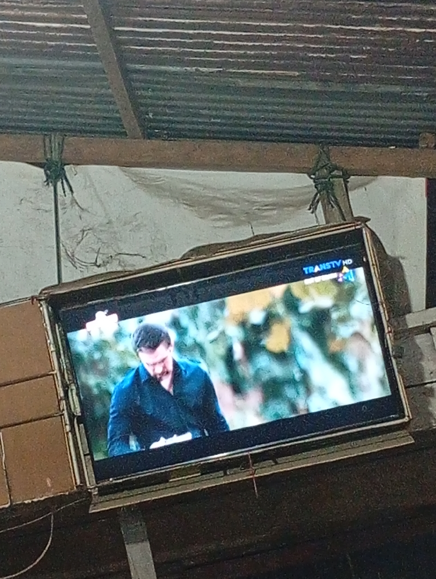 The image shows a television screen displaying a man wearing a dark blue shirt. The TV is mounted outdoors, likely under a corrugated metal roof supported by wooden beams. There is a white tarpaulin behind the television, attached with rope to the beams. The scene suggests an informal outdoor setup, potentially in a residential or community area. The overall impression is one of a simple, rustic environment. The specific location in Lhokseumawe, Indonesia, is not visually identifiable from the image itself.