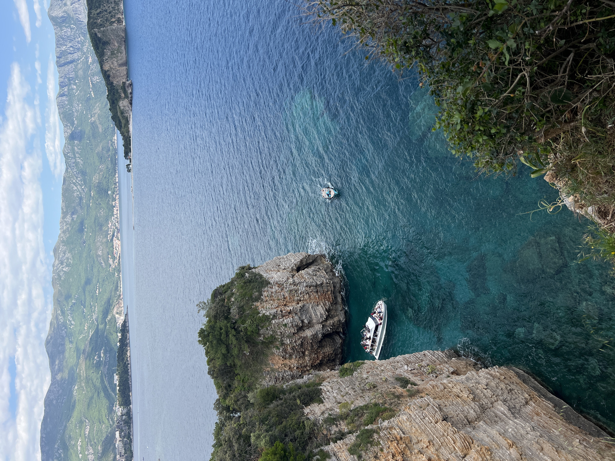 View from above of the Adriatic Sea, with a rocky coastline and mountains in the distance in the city of Budva, Montenegro.
A white motorboat filled with people sits next to a rocky cliff face with green vegetation. A smaller boat is visible further away. The water is clear and turquoise in color near the cliffs, fading to a deeper blue in the distance. Vegetation grows on the top of the cliffs and hangs over the edge. The cliffs are layered with horizontal lines that indicate rock formations. Mountains and a narrow strip of land can be seen in the background. The sky is clear.