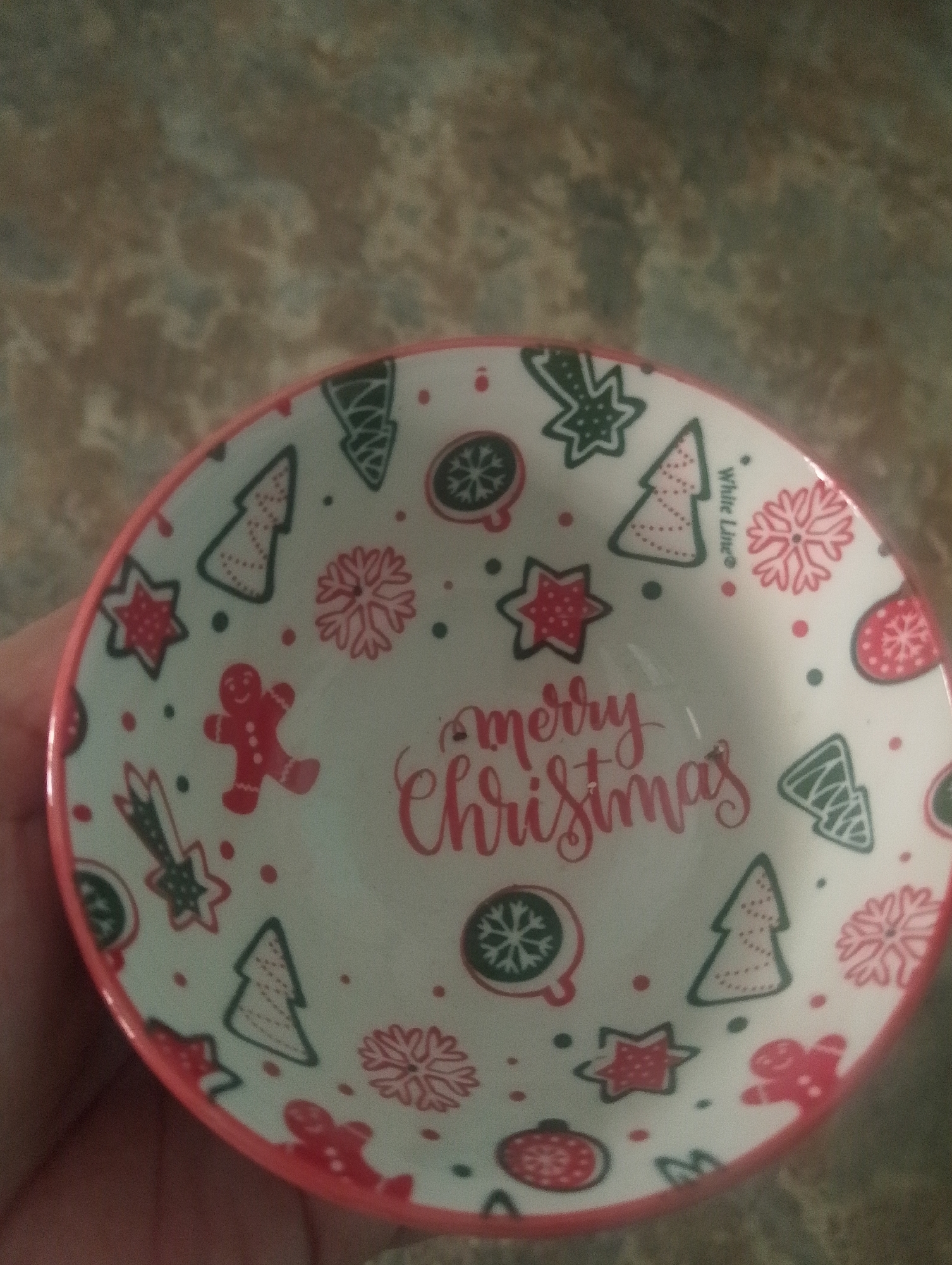 A white, circular bowl with a red rim is decorated with Christmas-themed imagery. The bowl is adorned with various designs, including gingerbread men, Christmas trees, snowflakes, stars, ornaments, and comets, all rendered in red and green outlines. In the center of the bowl, the words "Merry Christmas" are written in a cursive red font. The bowl is held by a person's hand, partially visible on the left side of the frame. The background is a textured surface, likely a countertop, with a marbled pattern of brown, gray, and beige hues. The image was taken indoors, with the lighting suggesting it was captured in a home environment. There is no information available about the location in Serbelawan, Indonesia.