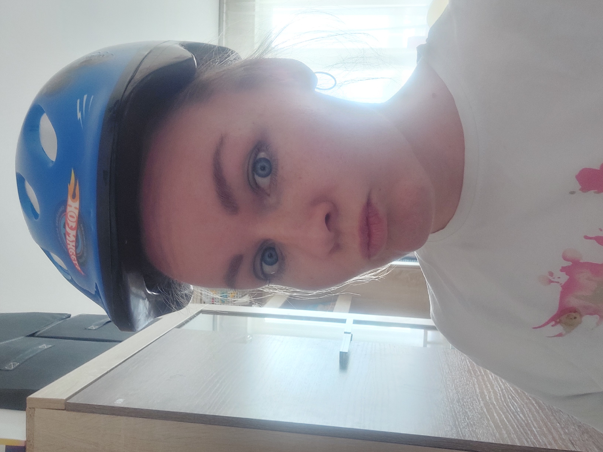 A woman wearing a blue Hot Wheels helmet and a white t-shirt with pink stains is posing for a selfie. The background appears to be indoors, with a window and furniture visible. There is no information to verify that the location is in the city of Chaikovsky, Russia.