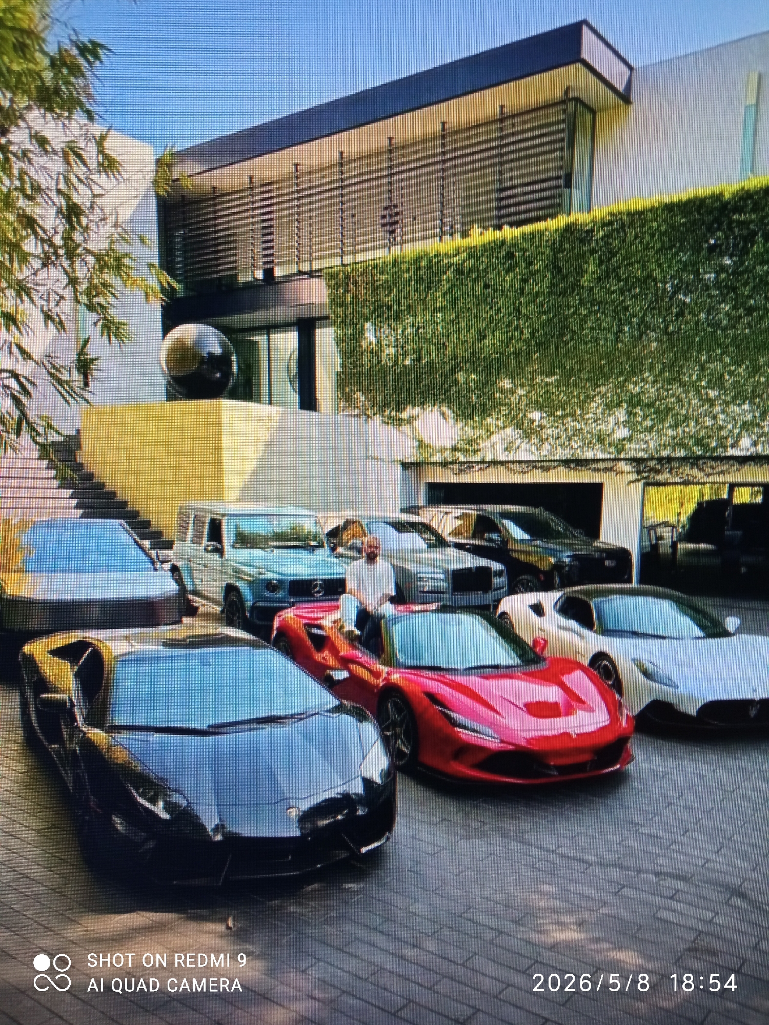 The image shows a large, modern house with multiple luxury cars parked in front of it. The house has a white facade, horizontal slats on the second floor, and a green hedge covering part of the building. There are stairs leading up to a yellow platform, and a black spherical sculpture is visible. The cars include a black Lamborghini, a red Ferrari, a white sports car, a light blue G-Wagon, and a Rolls Royce. Further back, there are two black SUVs parked inside an open garage. A man in a white shirt is standing near the Rolls Royce. The timestamp on the image indicates a date of May 8, 2026, at 18:54. The location is listed as Unknown, Unknown.