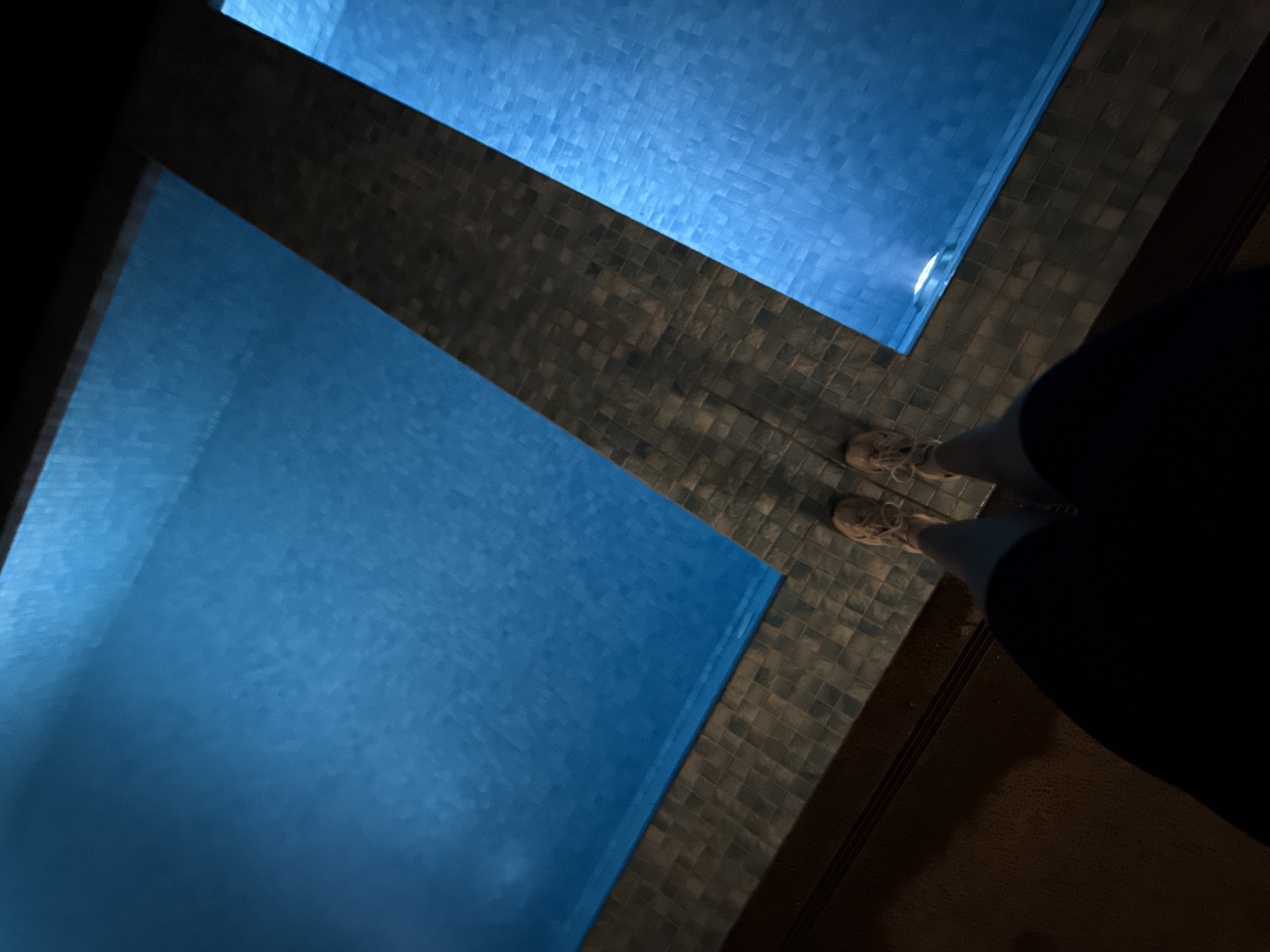 An overhead shot depicts two pools filled with blue water, separated by a tiled walkway. A person stands on the tiled walkway, wearing shoes and dark clothing. The photograph was taken indoors. Location: Jinhua, China.