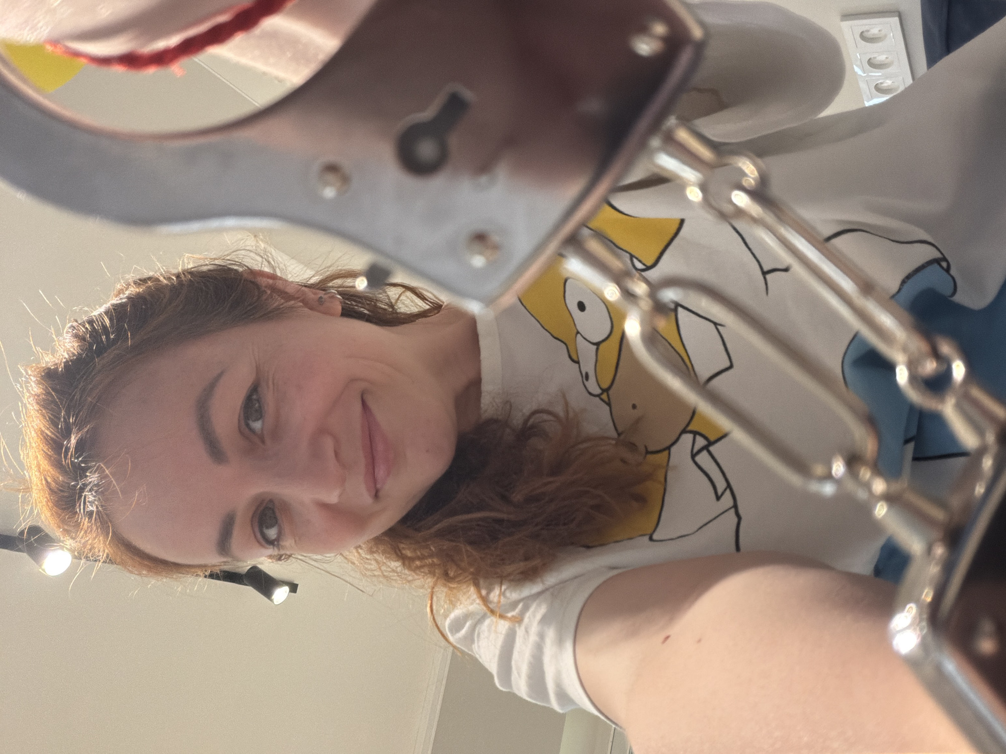 A woman is photographed from below, likely by herself. She has light skin, reddish-brown hair, and is smiling. She wears a white t-shirt with a yellow cartoon character print, possibly Homer Simpson. The photograph is taken through or with a pair of silver-colored handcuffs, which are in the immediate foreground and slightly out of focus. The location appears to be indoors, illuminated by a track lighting system. No specific details identify the city of Chaikovsky, Russia.