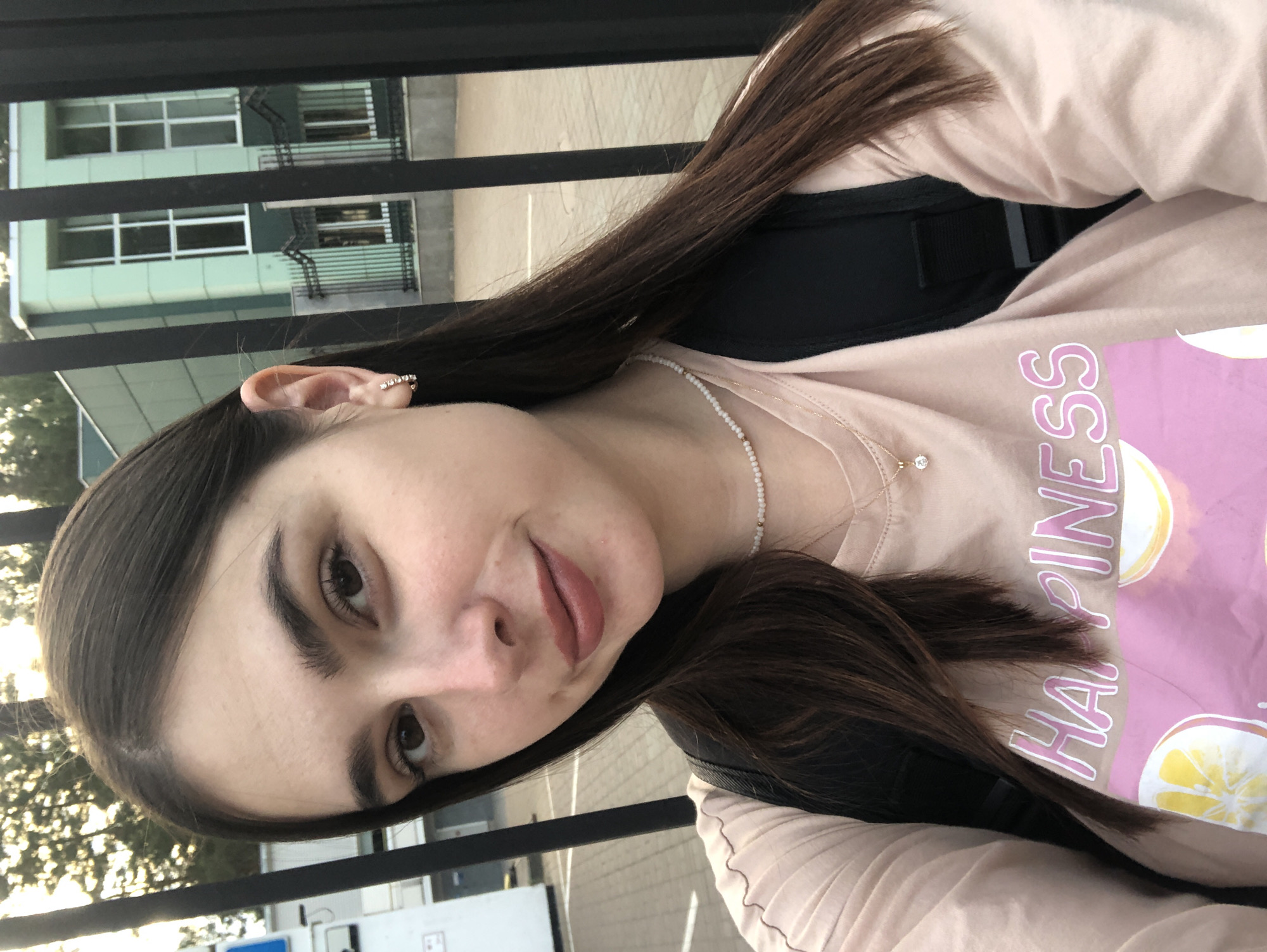 A young woman with long, dark brown hair and brown eyes is pictured in a selfie. She is wearing a pale pink t-shirt with the word "HAPPINESS" printed on it in a pink and white font, along with a graphic of sliced lemons and oranges. She has a delicate pearl necklace and a gold chain with a small diamond pendant. Her left ear is adorned with a hoop earring featuring small, clear stones. She appears to be standing outdoors behind a black metal fence, with a light green building visible in the background, possibly a school or administrative building. The environment suggests an urban or suburban setting. The lighting indicates it is daytime, possibly late afternoon. The overall scene is casual and friendly.