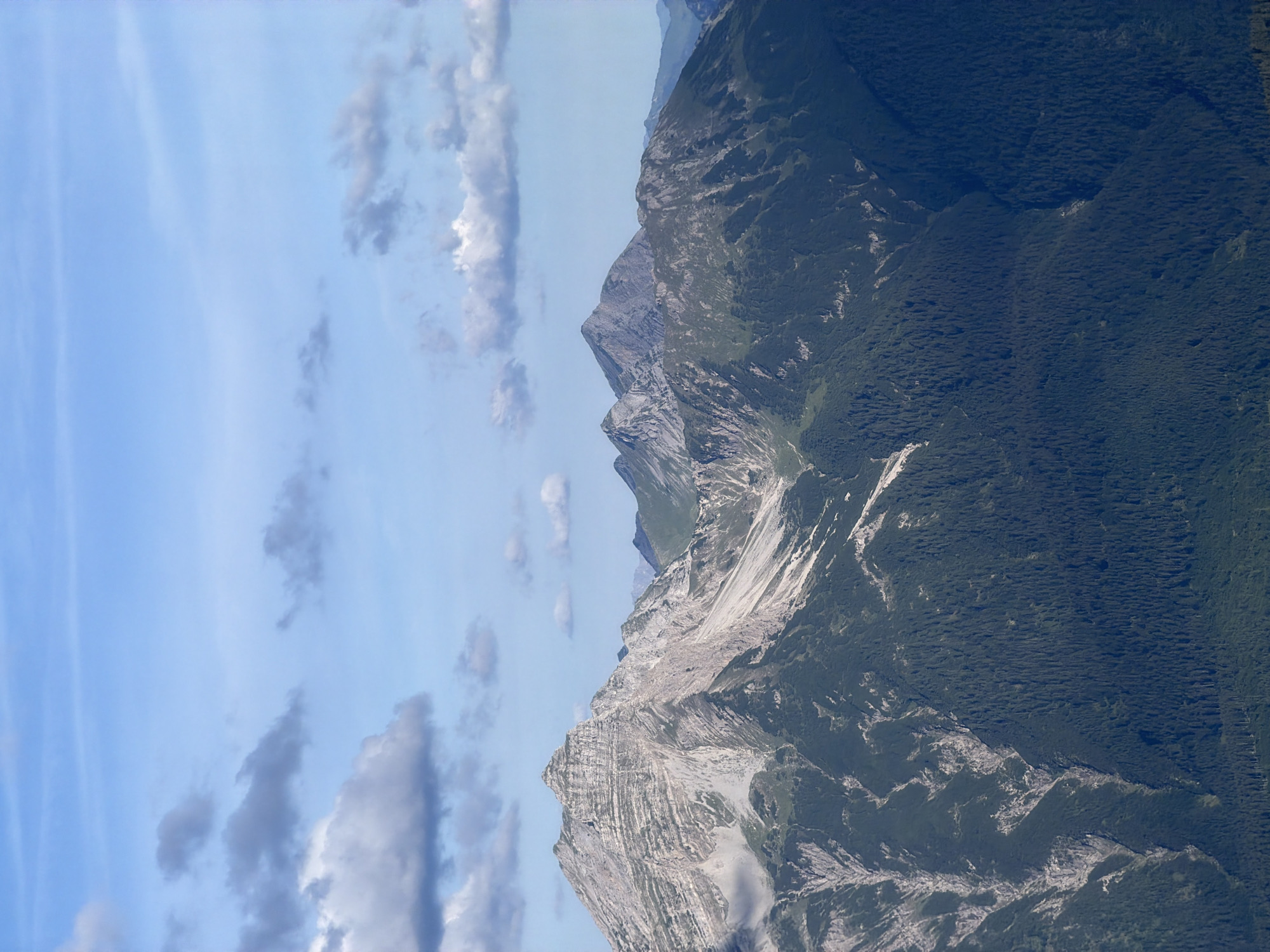The image displays a mountain range under a partly cloudy blue sky. The foreground is dominated by dark green, densely packed coniferous trees that cover the lower slopes of the mountains. Above the treeline, the mountains transition to rocky, barren, and scree-covered slopes with visible geological strata. Several peaks are visible, with the most prominent one in the left-center of the frame exhibiting steep, textured cliffs and ridges. In the background, further mountain peaks recede into the distance, some appearing darker and more rugged. The sky is a light blue with scattered white and gray clouds, some of which are wispy and stretched horizontally, while others are more cumulus-like. The overall impression is a vast, natural, and rugged mountain landscape. The image does not contain any people or man-made objects other than what may be inferred as trails or geological features on the mountain slopes.
While the image depicts a mountain landscape, it does not specifically show or indicate the location of Grainau, Germany. The provided image is a general mountain scene.