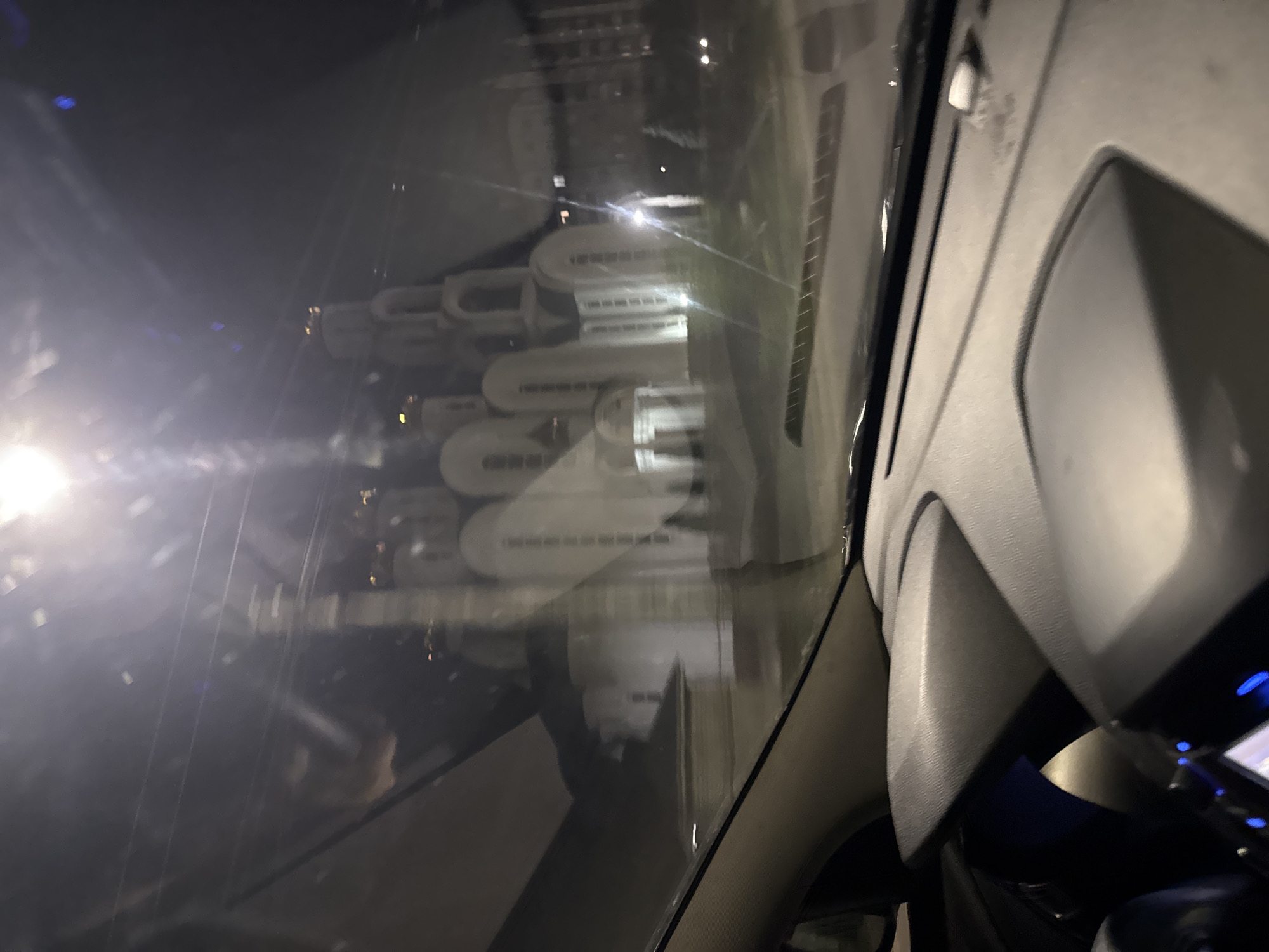 The image is a nighttime, interior shot from a vehicle, looking through the windshield. A large, white building with multiple domed roofs, likely a church or cathedral, is visible in the background. The building is illuminated, and its architectural features suggest Eastern Orthodox design. There are lights within the building and around its exterior. The foreground shows the interior of the car, with the dashboard and part of the steering wheel visible. The overall scene is slightly blurry due to the low light conditions and possibly motion.
This structure is identified as the Cathedral of the Nativity of the Blessed Virgin Mary, located in Rostov-on-Don, Russia.