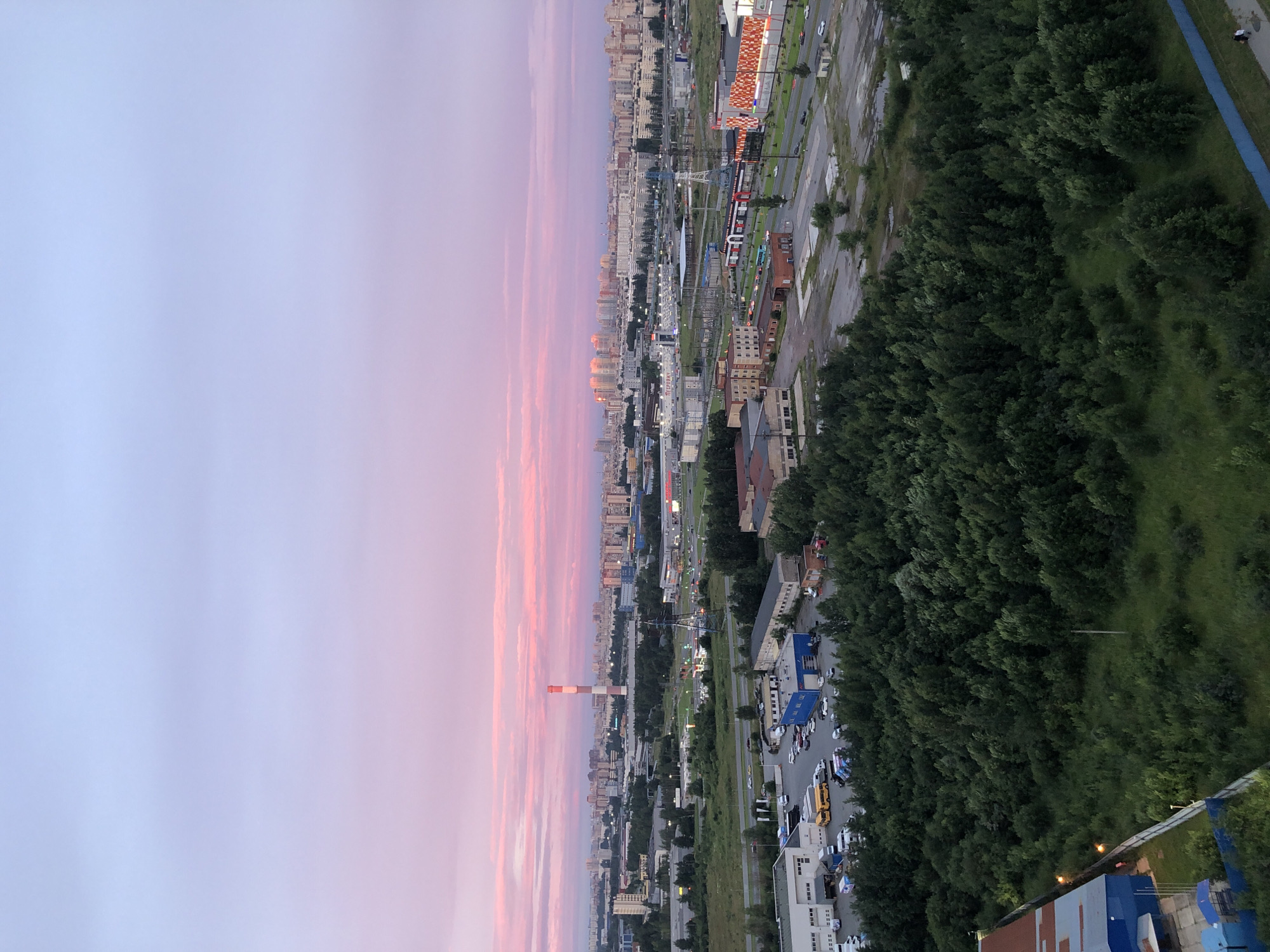 This is a high-angle, panoramic, medium shot of an industrial and residential area at dusk. The sky transitions from a pale blue at the top to soft pinks and purples near the horizon, with wispy clouds. In the distance, a city skyline with numerous buildings is visible. Closer to the foreground, there are industrial buildings, including a tall, red and white striped smokestack. A dense area of green trees occupies the mid-ground, with a few roads and parking lots interspersed. Several vehicles, including cars and buses, are parked or driving on the roads. In the lower left corner, a portion of a blue industrial building with some exterior lighting is visible. The overall scene suggests an urban environment with both industrial and residential elements, captured during the twilight hours.