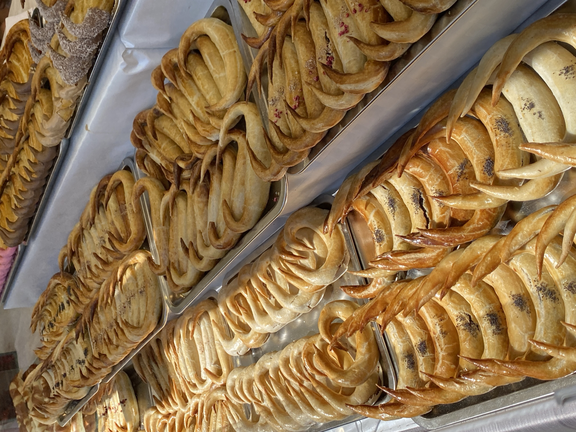 The image shows a selection of freshly baked pastries displayed on metal trays. The pastries appear to be crescent-shaped with various toppings, including seeds and powdered sugar. The location is likely Budapest, Hungary, as these types of pastries are common in Hungarian cuisine. The pastries are arranged in rows, suggesting they are available for sale.