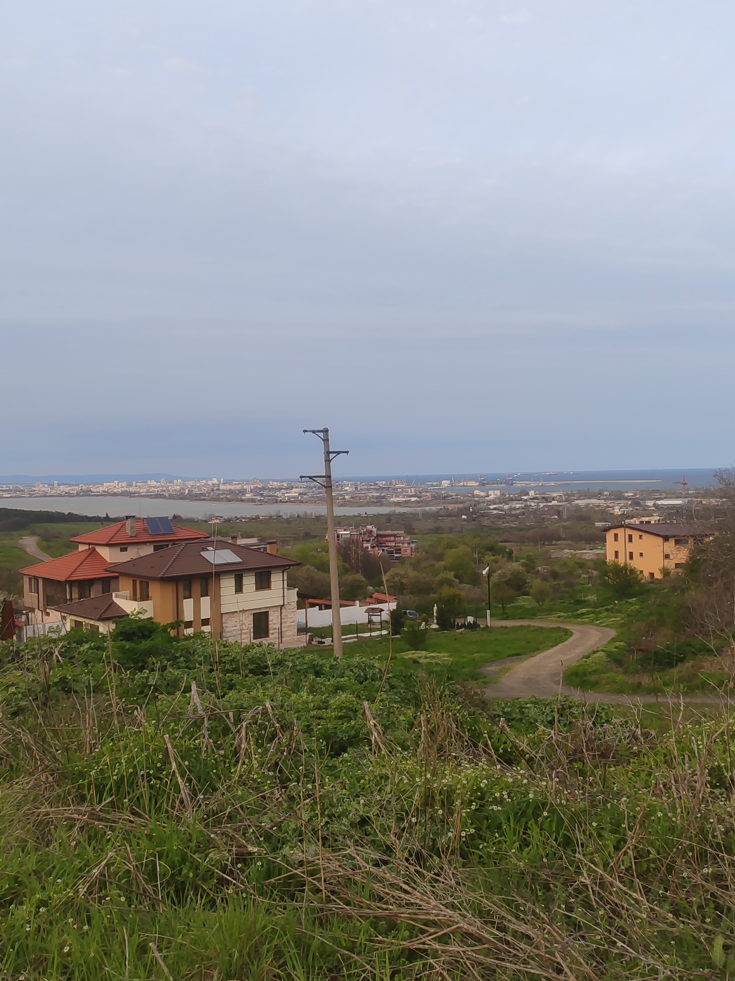 The image depicts a landscape overlooking a cityscape, likely Burgas, Bulgaria, based on the geography. In the foreground, there is abundant green vegetation and dried brown vegetation. Several residential buildings with red and brown tiled roofs are visible. A dirt road winds through the landscape towards the city. In the distance, a body of water separates the foreground from the dense urban area. Utility poles are visible, and the sky is overcast. The overall setting suggests a vantage point on the outskirts of the city, offering a panoramic view.