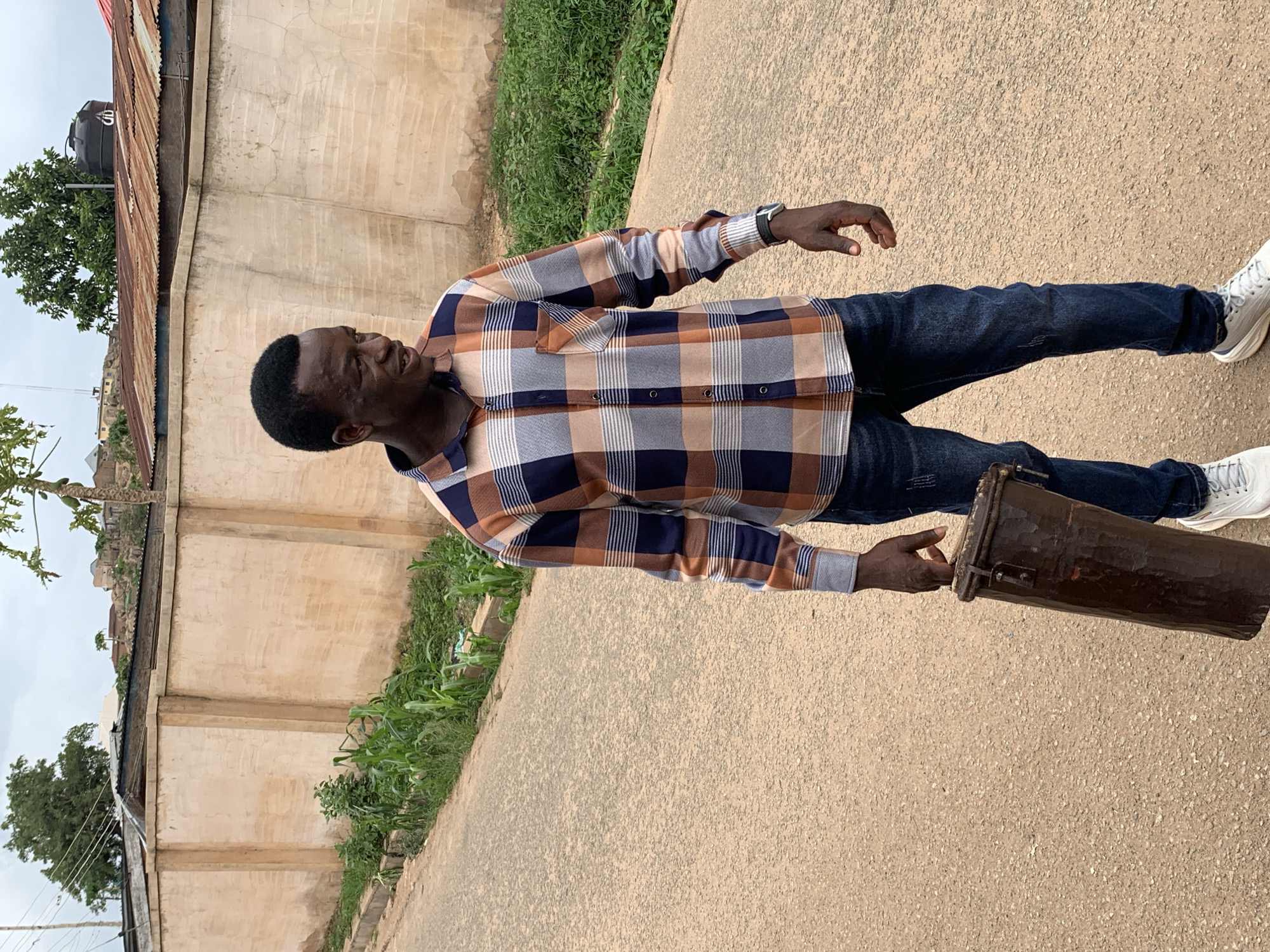 An African man wearing a long-sleeved plaid shirt and dark jeans stands on a paved surface. He is holding a brown cylindrical object, possibly a drum, in his right hand. He has a dark complexion and short hair. He is wearing white sneakers and a watch. Behind him is a concrete wall with some vegetation growing near it. In the background, there is a building with a corrugated metal roof and a water tank. The scene appears to be located in the city of Jos, Nigeria. The weather is overcast.