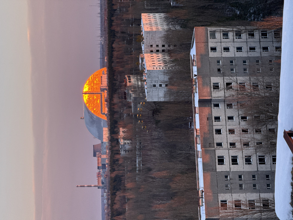 An aerial or elevated perspective captures a desolate urban landscape in Prypiat, Ukraine, at either sunrise or sunset. The sky displays hues of pink and light purple. In the foreground, a snow-covered surface precedes a multi-story, grey concrete residential building with numerous empty window frames, reflecting orange light on its upper facade. Behind this, a dense, leafless forest spans the mid-ground, interspersed with other abandoned, multi-story concrete buildings. Among the trees, the large, rusted Ferris wheel of the Prypiat amusement park is visible. Further in the background, beyond the forest line, the industrial structures of the Chernobyl Nuclear Power Plant are present. The most prominent feature is the New Safe Confinement (NSC) arch, a large, semi-cylindrical structure covering the damaged Reactor 4, with its western facade intensely illuminated in bright orange by the low-angle sunlight. To the left of the NSC, a tall, slender industrial chimney and other power plant buildings are discernible. No human figures or active vehicles are visible.