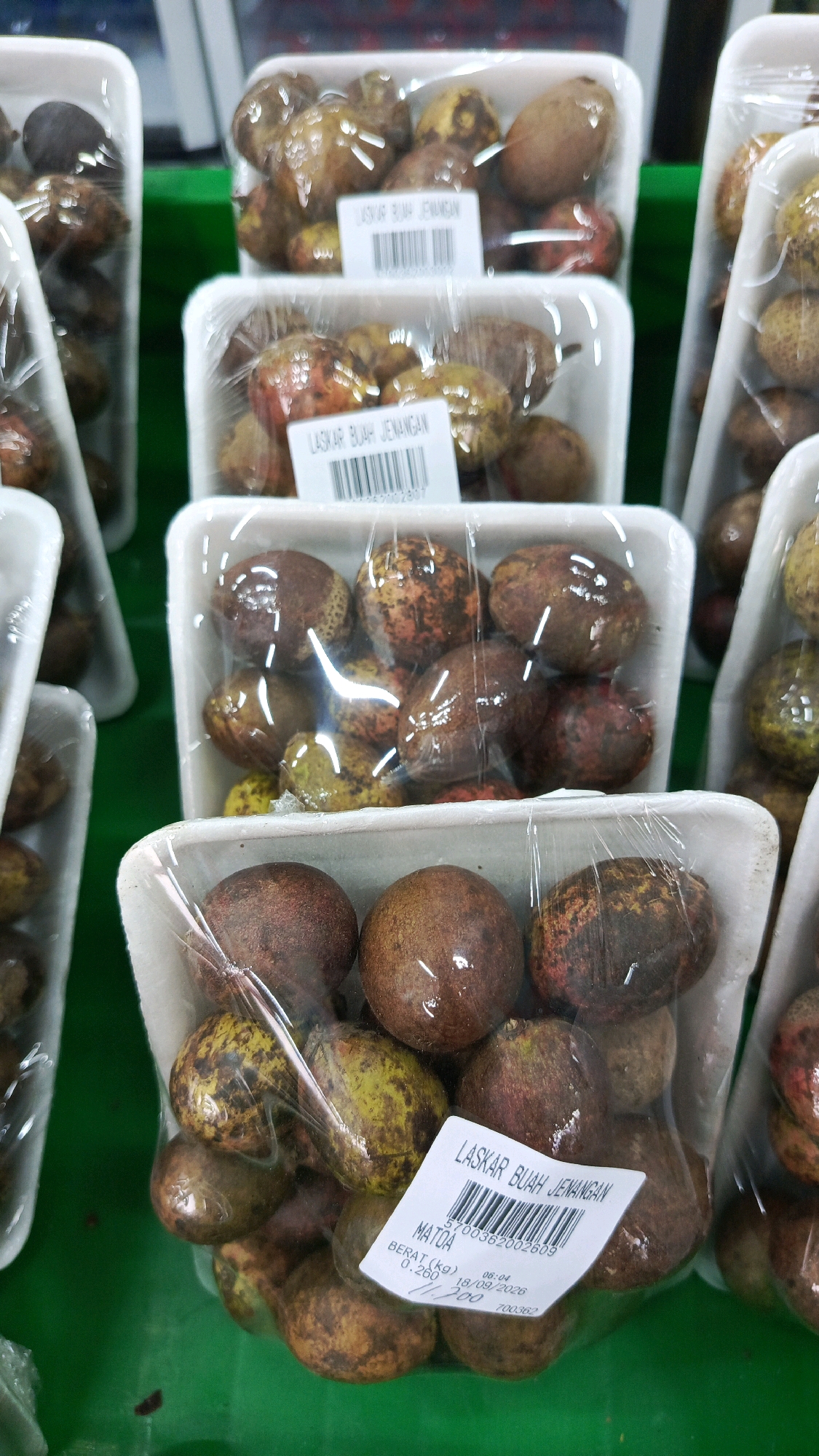 The image displays several plastic-wrapped containers of fruit, likely sold in a market or grocery store. The containers are white styrofoam and are stacked in multiple rows. The fruit inside appears to be small, round, and has a mottled brown and yellow/green skin. Some containers have labels with text that includes "LASKAR BUAH JENANGAN", "MATOA", "BERAT (kg)", and a date of "18/09/2026". The overall setting suggests a commercial display of produce. The background is blurred but appears to be a green surface. The lighting is bright, creating reflections on the plastic wrap. There are no people visible in the image.