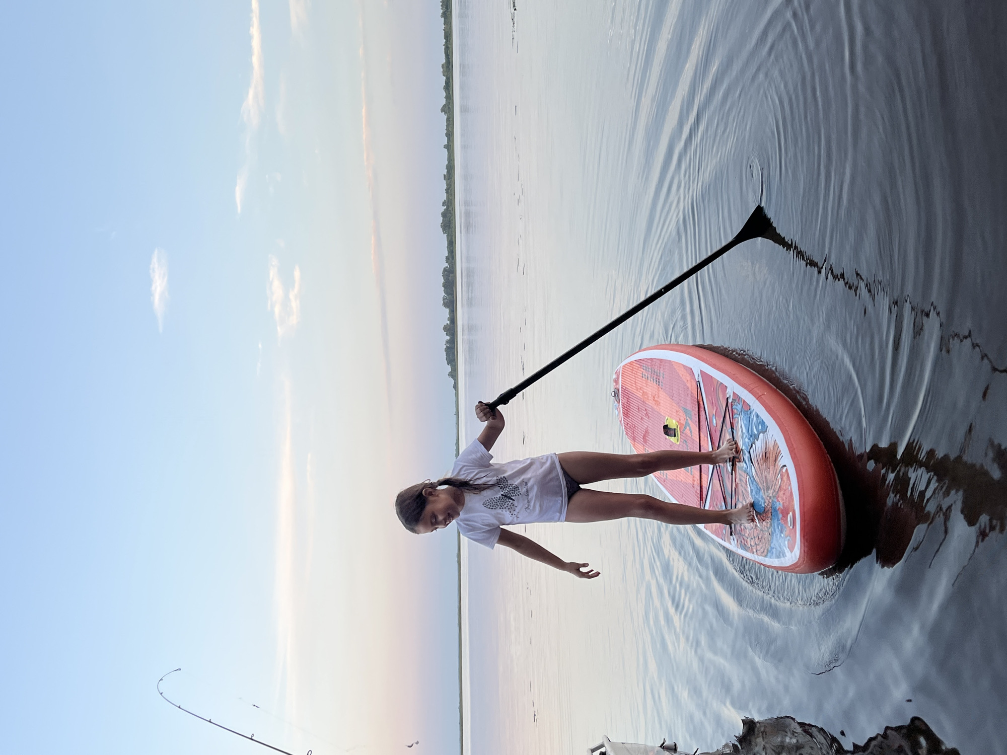 A young girl, dressed in a white t-shirt and dark bottoms, stands on an orange and white paddleboard in a calm body of water. She is holding a black paddle, with the blade submerged in the water. The girl is looking down and to her left. The paddleboard is decorated with a red and blue design. In the background, the sky is a gradient of pale blue and pink, with a thin line of clouds on the horizon. A fishing rod is visible on the left side of the image, extending from the left edge upwards and curving towards the right. The water is calm, with subtle ripples and reflections. The shoreline is visible in the far distance as a line of trees. This scene is taking place at dusk in Ust-Belsk, Russia.