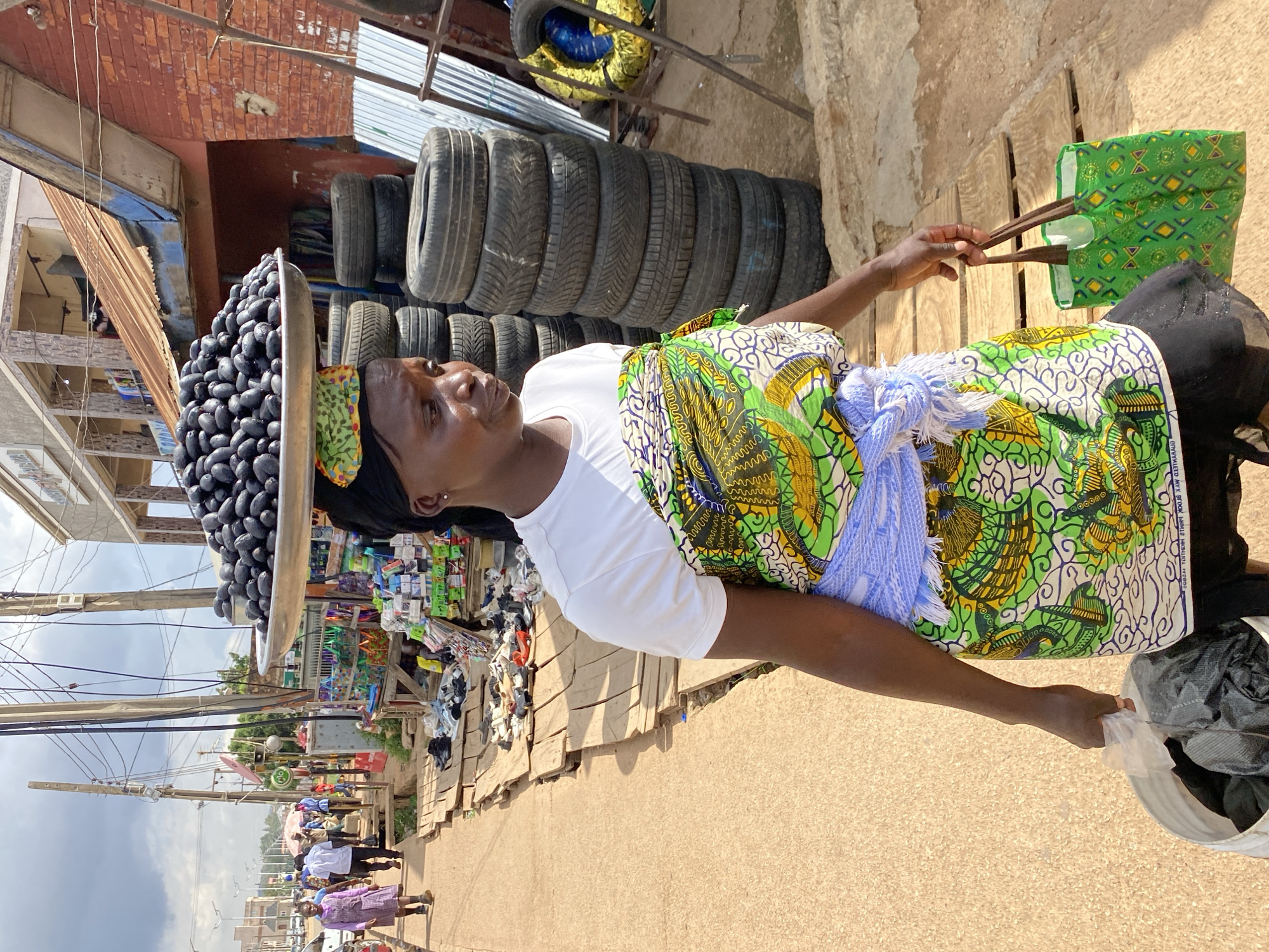 A woman is walking on a wooden plank path in a marketplace in Bukuru, Nigeria. She is carrying a large metal tray of dark, plum-like fruits on her head, balanced by a patterned cloth. She is wearing a white t-shirt and a brightly colored green and yellow patterned wrap skirt, with a blue and white striped sash around her waist. In her right hand, she carries a green patterned tote bag. In her left hand, she holds a plastic bag filled with dark objects, which she is carrying in a white bucket. Behind her, stacked tires are visible, along with market stalls selling various goods. People are walking in the background on a sandy path. The sky is overcast.