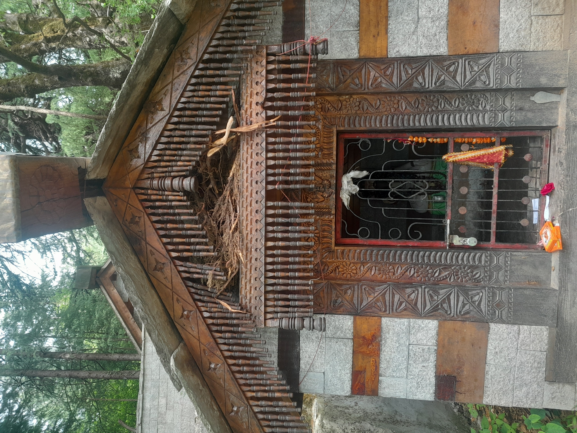 The image shows the entrance to a small shrine or temple building, likely in a rural or mountainous area, possibly in the Himalayas given the architecture and surrounding vegetation. The building features a stone facade with alternating horizontal bands of gray stone and reddish-brown wood. Above the entrance, there is an elaborately carved wooden roofline with decorative elements and dangling wooden spindles. A portion of the roof is covered in dried grass or reeds, possibly indicating a nest or recent disarray.
The entrance itself is a dark red metal gate with a padlock. Through the gate, a statue of a deity or revered figure is partially visible, draped in green cloth. Hanging from the gate are prayer flags and a string of marigold flowers. A small piece of white cloth is affixed to the inside of the gate.
To the left of the entrance, there are several scattered items on the ground, including a bright orange packet and a single red rose petal. The surrounding environment is lush with green foliage and tall trees, suggesting a natural setting. The overall impression is of a sacred place that is integrated into its natural surroundings and shows signs of both devotion and some level of natural weathering or disruption. The architectural style, particularly the wooden carvings, is indicative of Himalayan craftsmanship.