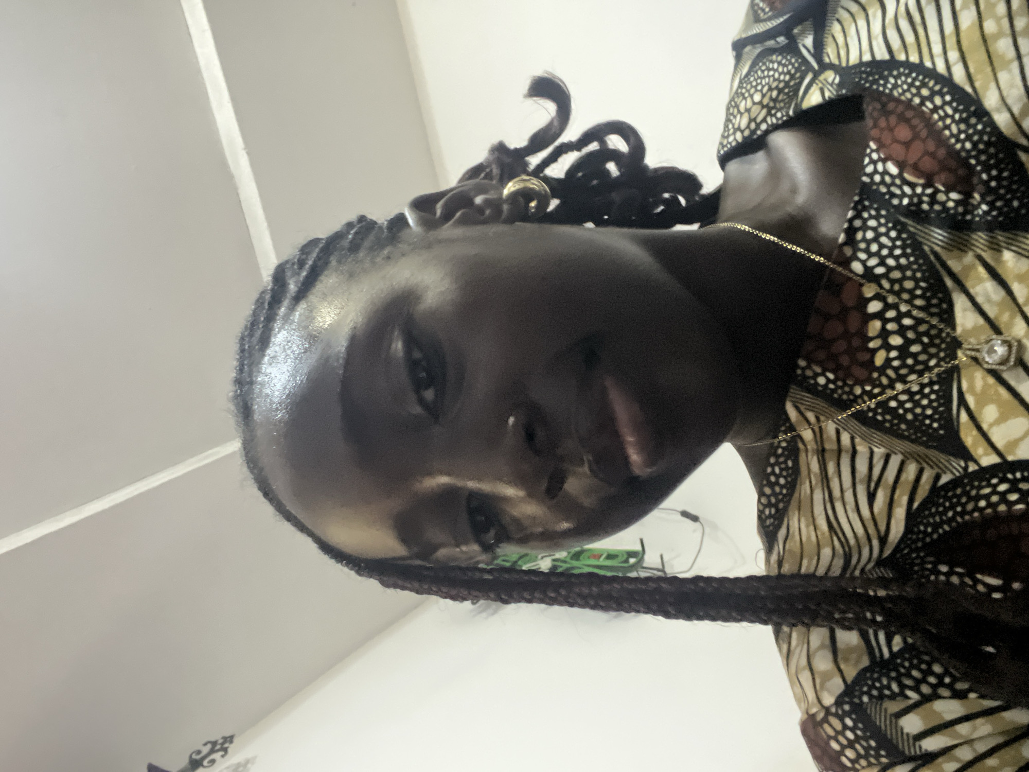 A medium-shot portrait features a young individual with dark skin and dark, thin braided hair, looking directly into the camera. Braids extend over the left shoulder, and some are styled in coils near the right ear. A prominent sheen is visible on the forehead and nose. The subject has full lips and a small nose piercing on the right nostril. A thick, gold-colored hoop earring is worn on the right ear, and a thin gold chain with a rectangular, clear gemstone pendant hangs around the neck. The individual is wearing a top or dress made of an Ankara-style fabric with an intricate pattern incorporating cream, black, brown, and reddish-brown colors with white accents.
The scene is an indoor environment in Abuja, Nigeria. The background consists of plain, light-colored walls and a matching ceiling with a visible seam. On the upper left wall, a dark, ornate decorative object is partially visible, along with a green object, possibly an electrical fixture or decoration, with associated wires. The lighting appears to be from an overhead source, casting a soft, even glow.