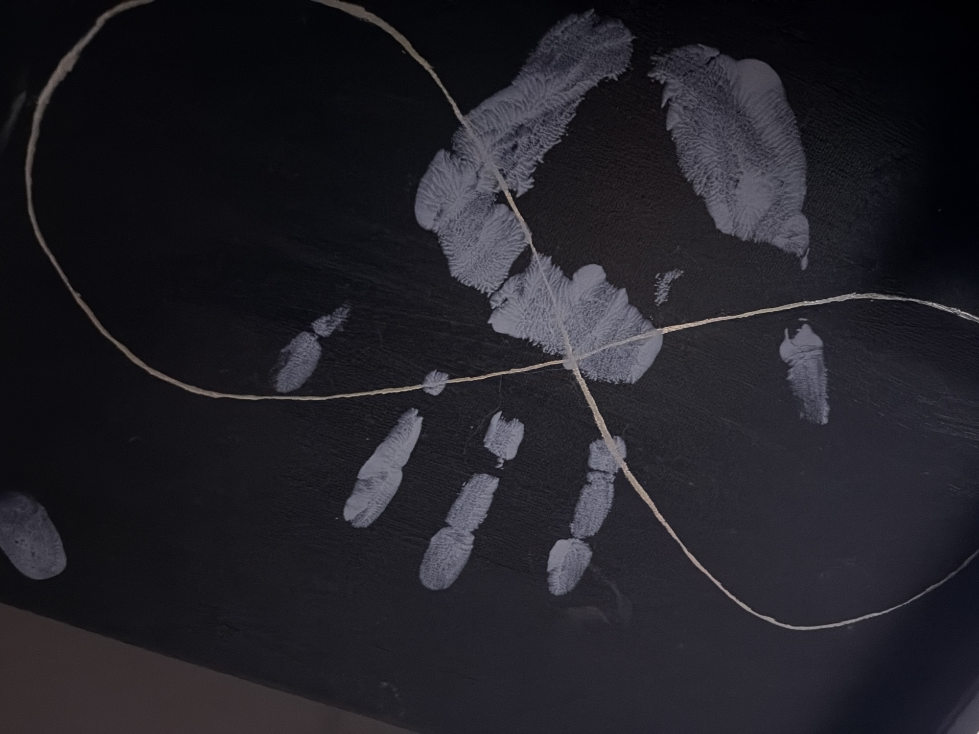 The image depicts a handprint with a piece of string or thread draped across it. The handprint appears to be made with a light-colored substance, possibly paint or ink, on a dark surface. The string is a light brown or tan color and forms an irregular loop that intersects the handprint. The overall impression is that of a still life or an artistic arrangement. There is no information available about the location in Amstelveen, Netherlands, or any forensic details related to people, subjects, objects, actions, or interactions.