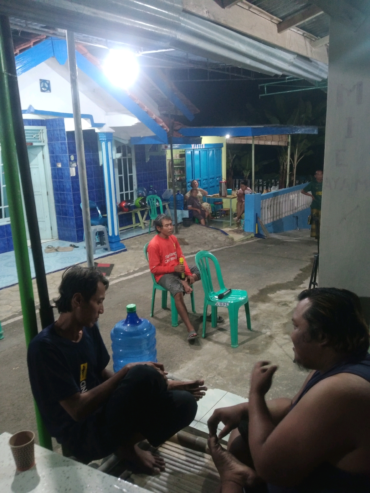 In Rejasari, Indonesia, at night, several individuals are gathered outdoors under a covered patio area. The scene suggests a casual social gathering. In the foreground, two men are seated on bamboo mats. One man, wearing a dark t-shirt and dark pants, is looking down. Next to him is a large blue water jug. To his right, another man, wearing a dark sleeveless shirt, is facing away from the camera, gesturing with his hands.
In the mid-ground, a man in an orange long-sleeved shirt sits on a green plastic chair, holding a yellow bottle. Another green plastic chair is nearby, with a microphone placed on it. Behind these individuals, a white house with blue trim is visible. In the background, other people are seated or standing, and there are more green chairs and tables. The environment appears to be an outdoor communal space, possibly a yard or a public area, with some vegetation visible in the distance. The lighting suggests artificial illumination from overhead fixtures.