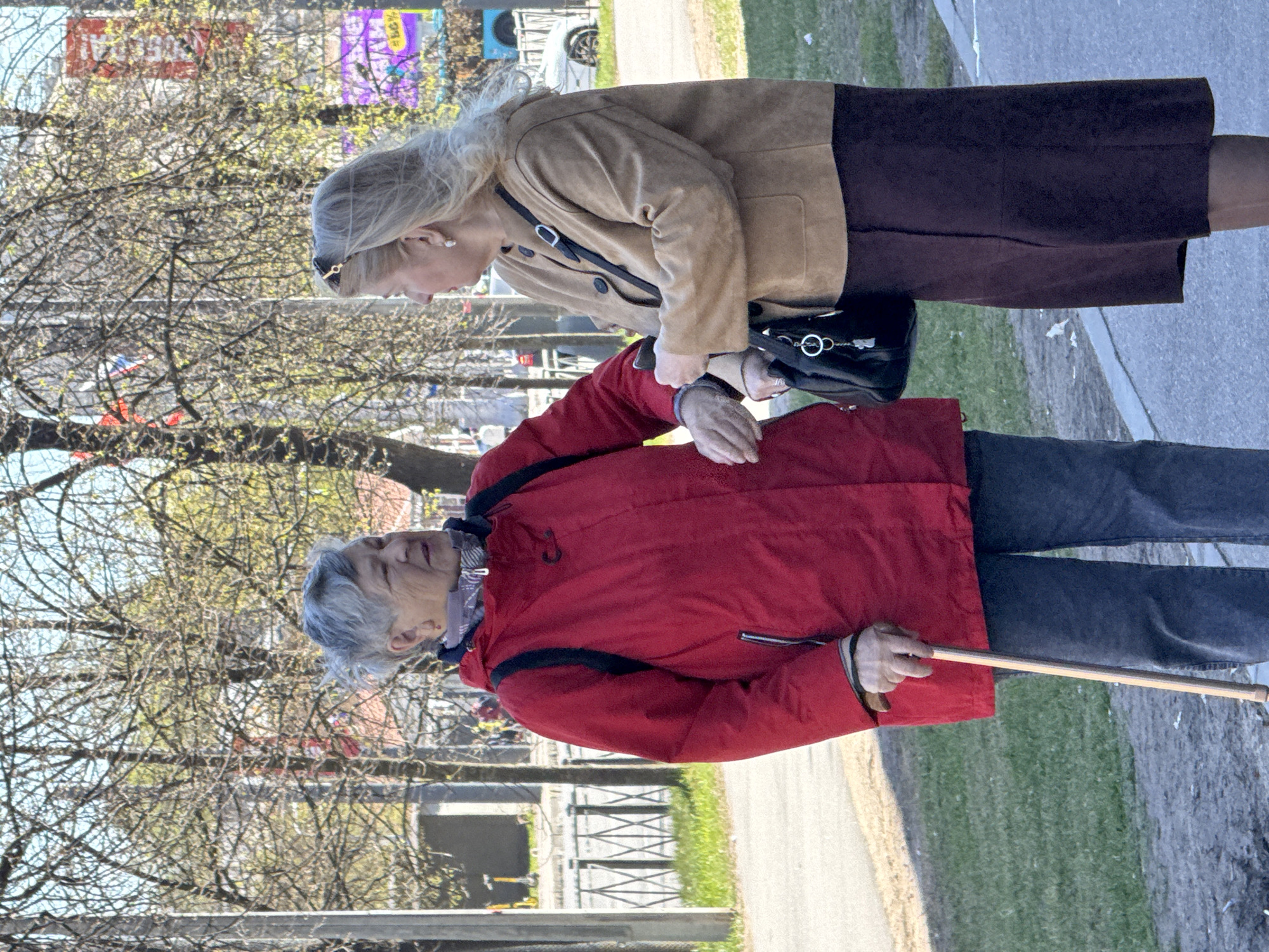 Two women stand on a paved walkway next to a grassy area and trees with bare branches. One woman, wearing a red jacket and gray pants, holds a cane. The other woman, wearing a tan jacket and brown skirt, looks at the woman with the cane and holds her wrist. The location is Saint Petersburg, Russia.