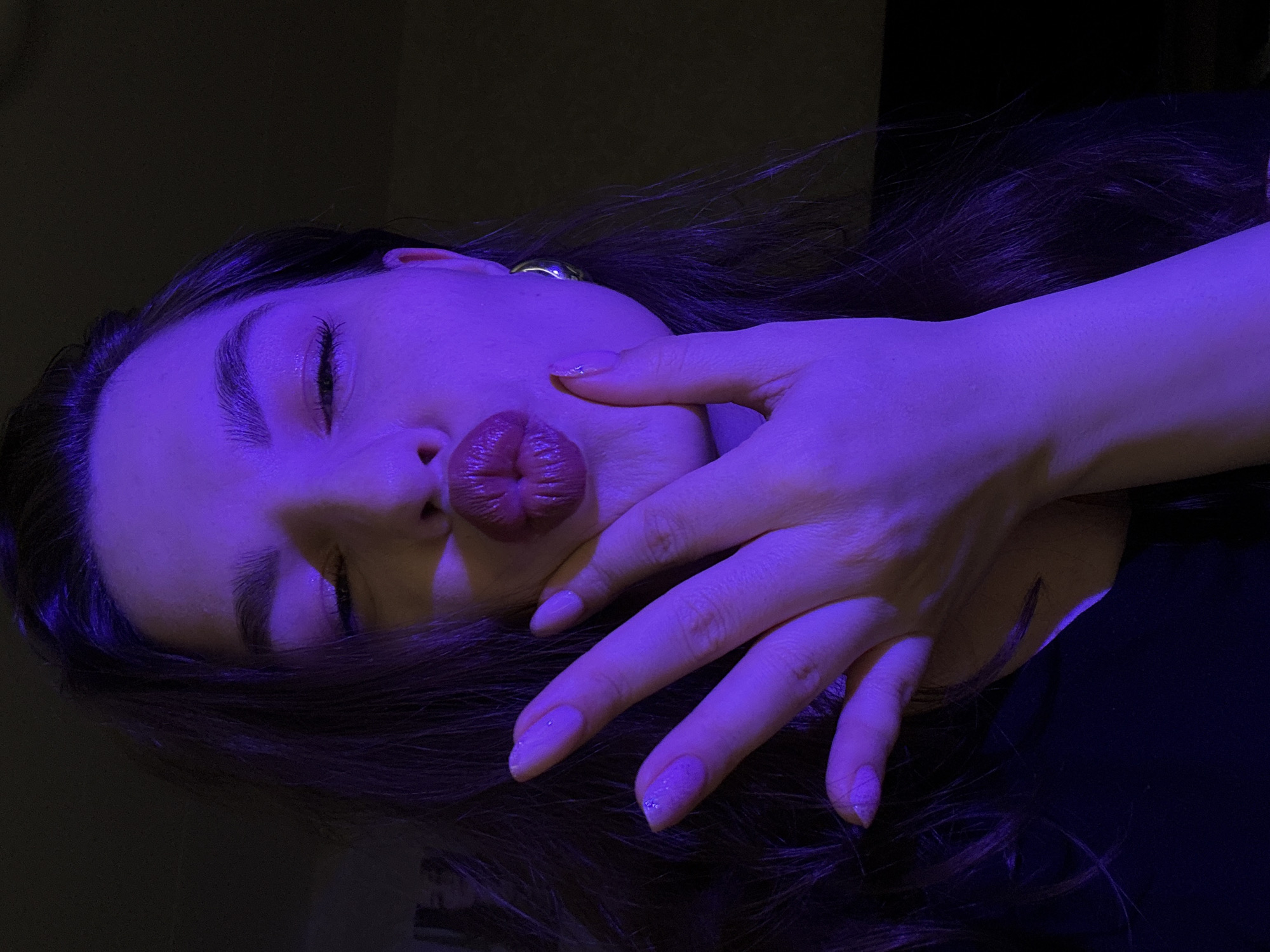 A close-up shot captures a young woman's face as she puckers her lips for a kiss, her left hand positioned near her face. The scene is illuminated by a purple light, casting a distinct hue across her skin and features. Her eyes are partially closed, and she wears light-colored eyeshadow and lip gloss. A silver earring is visible on her right ear. Her nails are painted a light purple color. The background is dark and out of focus, providing little environmental detail. The location in Chaikovsky, Russia is not discernible from the image.