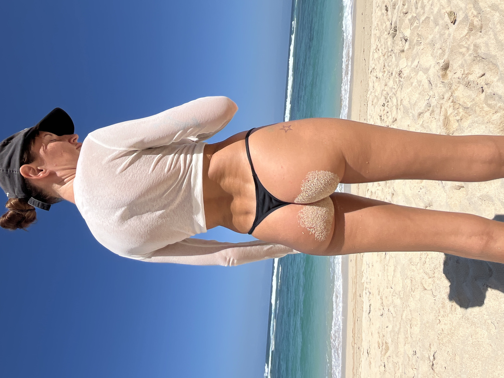 A woman stands on a sandy beach in Kuta Selatan, Indonesia, with her back to the camera. She wears a white long-sleeved cropped shirt and black thong bikini bottoms. Sand covers her buttocks. The ocean with waves and a clear blue sky are visible in the background.