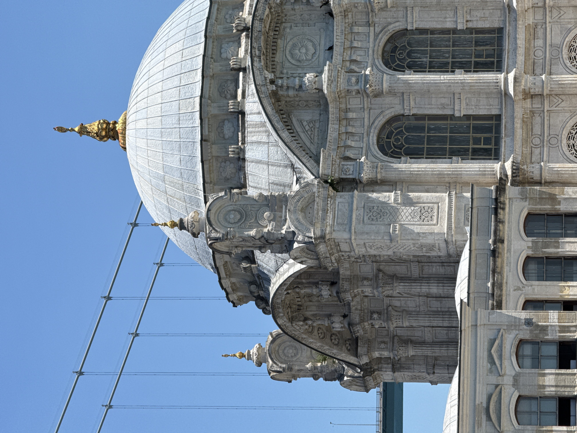 A close-up, eye-level shot captures the ornate stone architecture of a mosque in Beşiktaş, Turkey. The mosque features a large, ribbed dome topped with a golden finial, surrounded by smaller domes and towers. Intricate carvings adorn the facade, with arched windows and decorative stonework visible. In the background, the blue cables of a suspension bridge stretch across a clear blue sky. The overall scene is one of grand architectural detail set against a bright, sunny day.