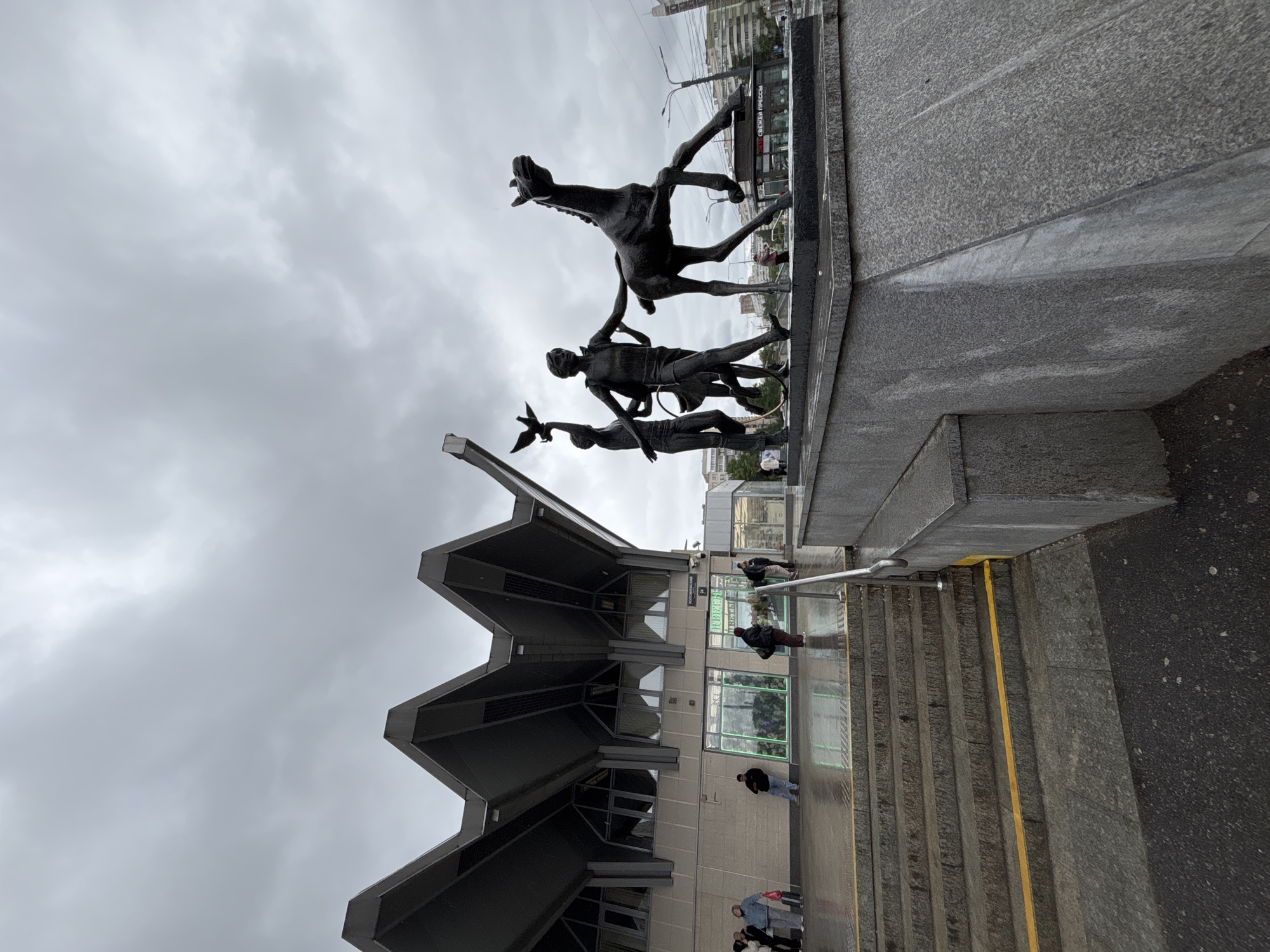 The image depicts a scene in Saint Petersburg, Russia. The focal point is a sculpture depicting a horse and figures. The sculpture is situated on a grey stone platform.
Behind the sculpture is a building with a sharply angled roof. People are visible near the entrance of the building. A staircase with a yellow stripe descends towards the building. The sky is overcast.