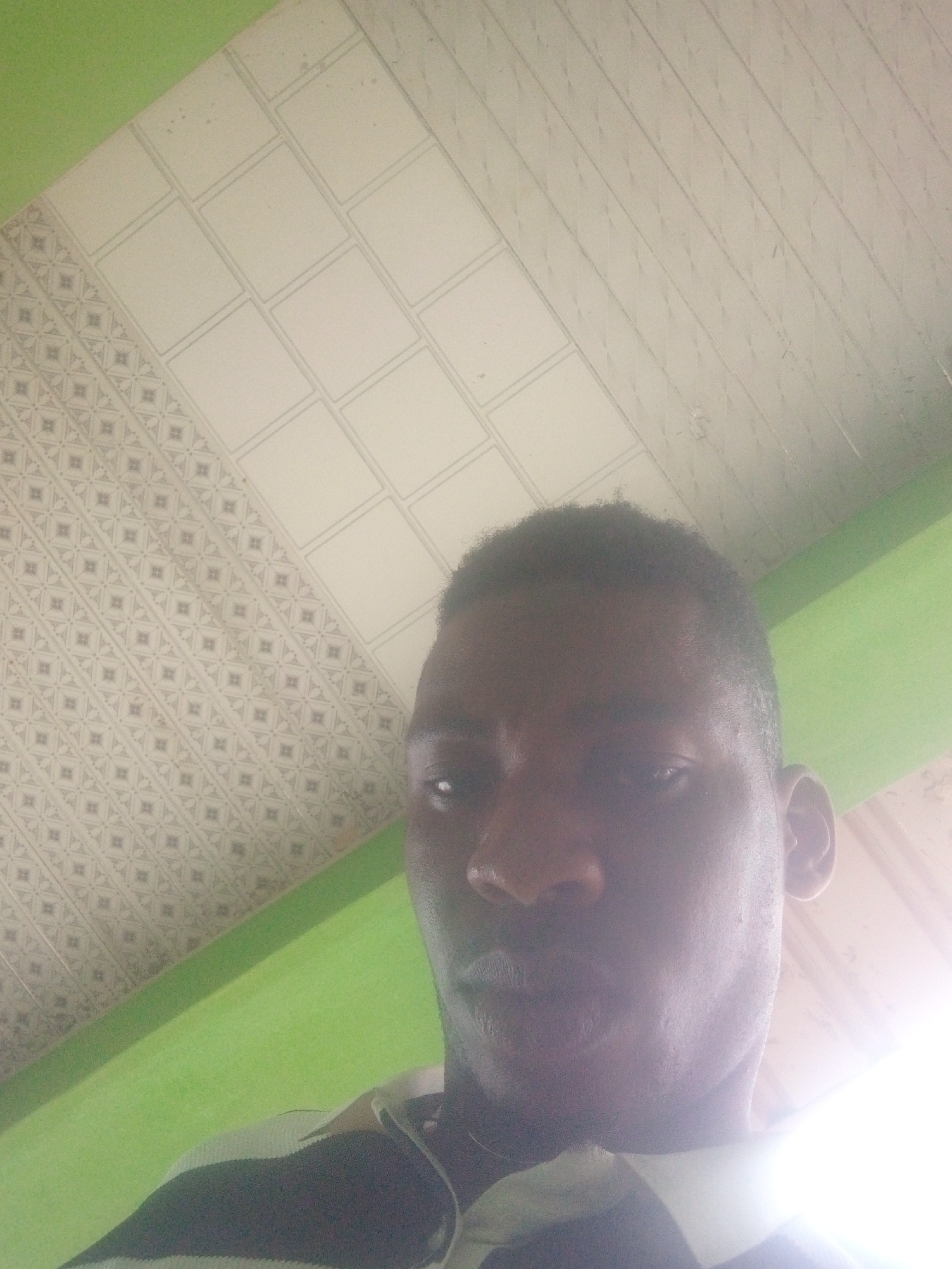 A close-up, upward-angled photograph shows a dark-skinned male with short hair, facing the camera. The man is wearing a striped shirt. Above him is a patterned ceiling, comprised of squares and small diamond patterns. A section of green wall is visible on the left side of the frame. The location is Unknown, Unknown.