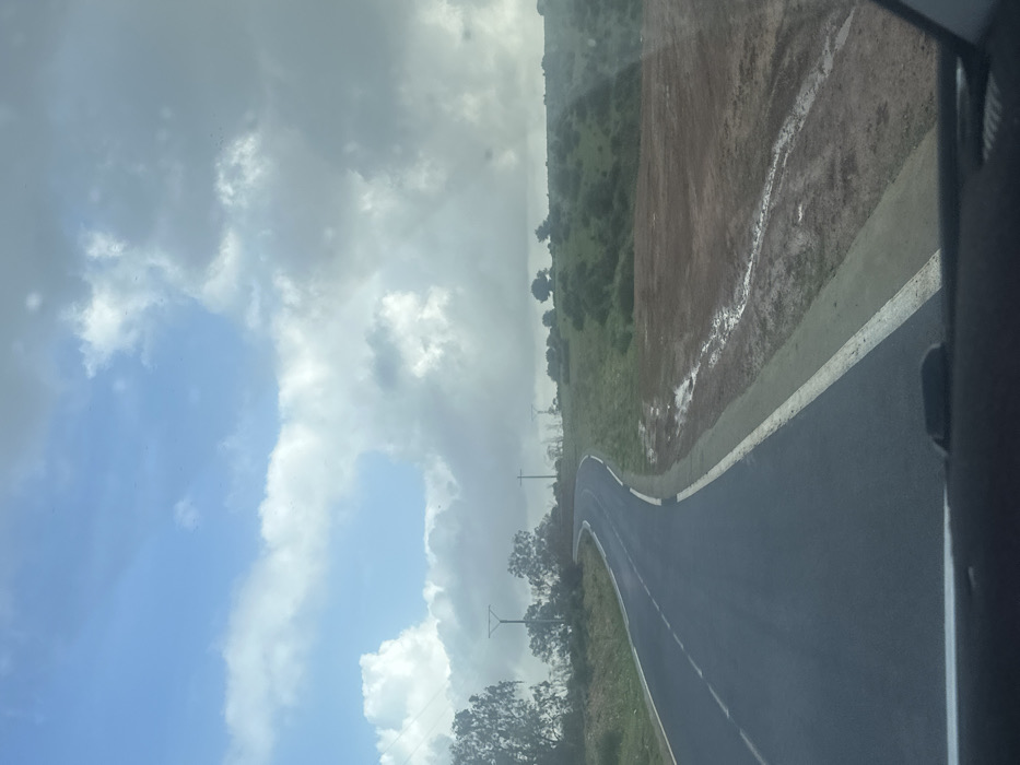 The image depicts a rural road scene in Oumazza, Morocco, viewed from inside a moving vehicle. A two-lane asphalt road, dark grey and appearing damp, curves gently left then right, receding into the mid-ground. It features a solid white line marking the right edge and a dashed white line in its center. To the immediate right of the road, a narrow grey shoulder borders a muddy, reddish-brown dirt strip exhibiting standing water and rivulets, indicative of recent precipitation. Beyond this, a gradual uphill slope is covered with green grass, scattered bushes, and small trees.
On the left side of the road, similar green vegetation and numerous trees line the route. Multiple utility poles, dark in color, are visible on both sides of the road in the mid-distance. The sky is partly cloudy; the upper left quadrant features clear blue sky with bright white cumulus clouds, while the upper right and central portions are dominated by larger, darker grey stratocumulus clouds. No individuals or animals are present.
