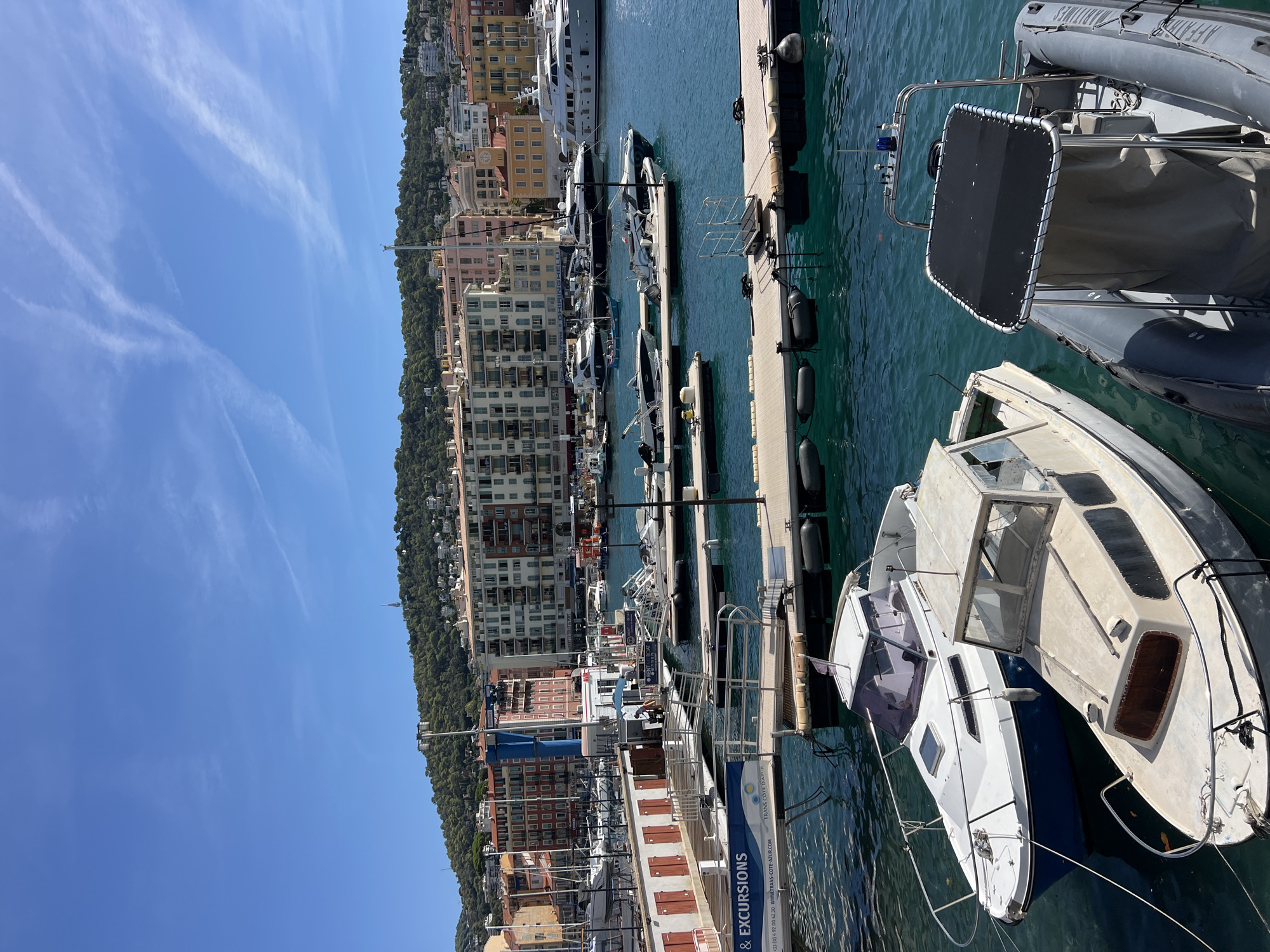The image depicts a sunny day at the port of Nice, France, known as Port Lympia. In the foreground, several boats are moored, including two smaller vessels with cabins, one white and blue, and another older, weathered cream-colored boat. To the right, a gray inflatable boat with a black canopy is visible. The middle ground features a series of floating docks extending into the turquoise water. Various other boats of different sizes are tied to these docks, with some larger yachts visible further in the background. A prominent sign on a walkway reads "RENT-A-BOAT". The background reveals the cityscape of Nice, characterized by a dense collection of multi-story buildings with colorful facades, typical of French Riviera architecture. Behind the buildings, a lush green hillside rises, dotted with more houses and topped with trees, under a bright blue sky with thin wispy clouds.
