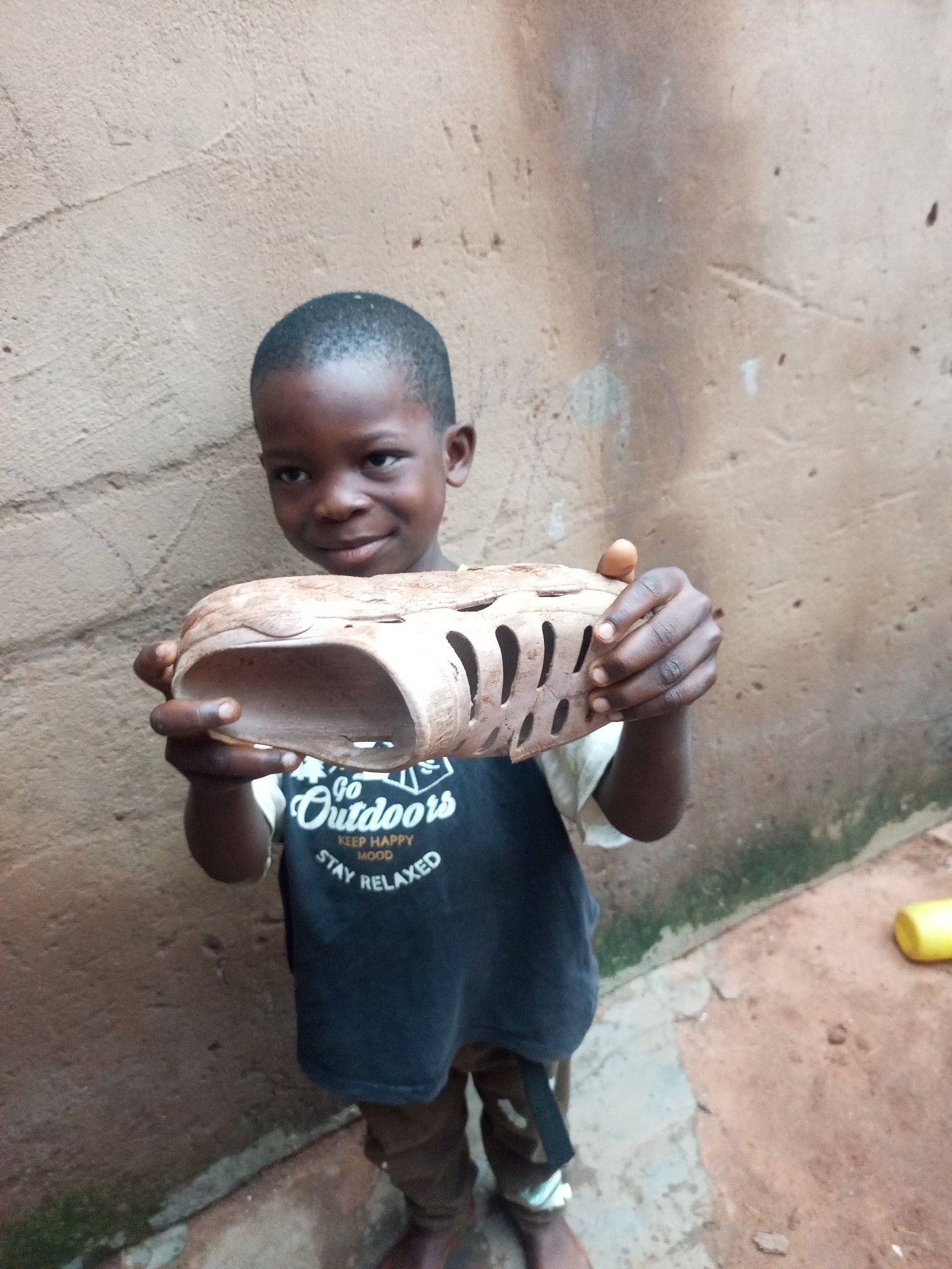 A young Black boy smiles as he holds a handmade sandal made from natural materials, possibly wood or dried plant matter, in front of a textured, light brown wall in Tempure, Nigeria. The boy wears a dark t-shirt with a white graphic that reads "Go Outdoors KEEP HAPPY MOOD STAY RELAXED" and brown pants. His feet are bare, and he stands on a dirt and stone surface. To the right, a bright yellow object, possibly a toy or tool, rests on the ground. The environment appears to be an outdoor, possibly rural, setting with the wall suggesting a building or enclosure.