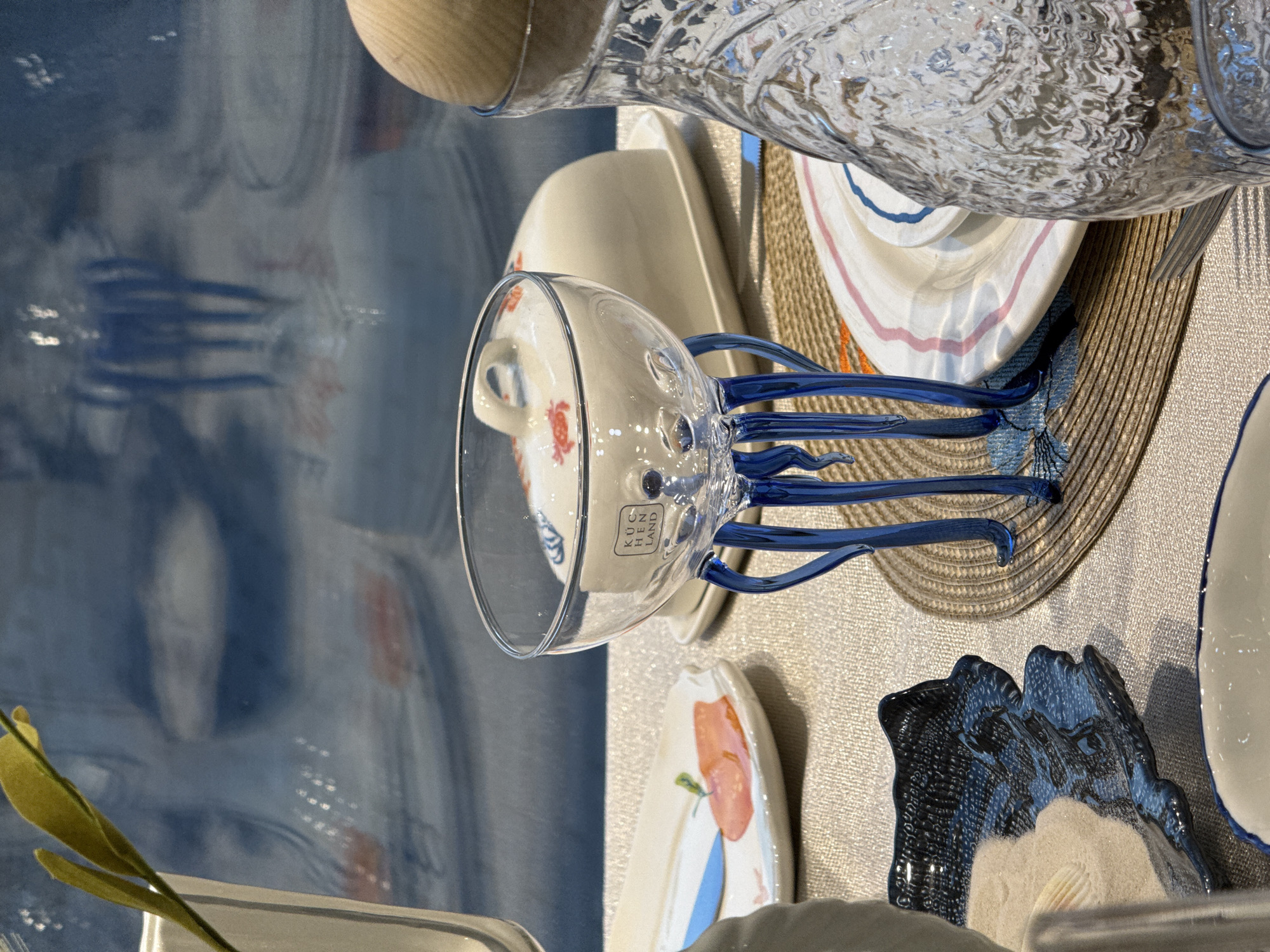 The image shows a display of dishware and glassware, arranged on a textured grey surface. Prominent items include a clear glass with multiple blue-colored leg-like appendages, a white plate with a red floral design, a blue decorative dish, a pitcher with a wooden lid, and a straw sun hat. The glass with the blue appendages has a small rectangular label that reads "KUCHEN LAND". The background suggests a reflective surface, perhaps a window or glass panel, with blurred light reflections and what appears to be a partial view of a city landscape. Given the "KUCHEN LAND" label and the presence of Russian letters on other items, this could be an exhibition or display within a store. The location is Krasnoyarsk, Russia.