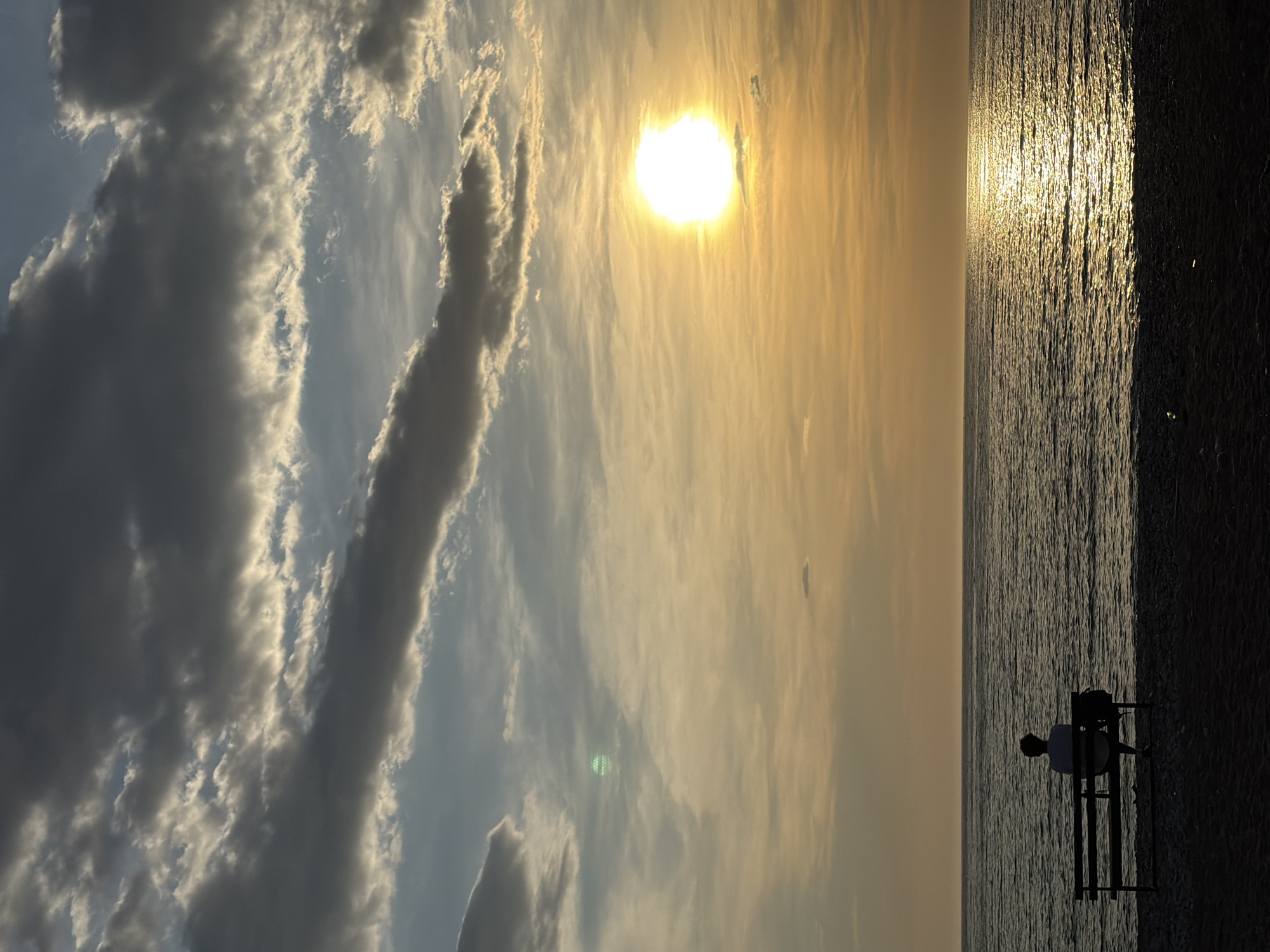 A landscape photo shows a beach scene at sunset in Bar, Montenegro. The sky, full of clouds, has an opening where the sun is visible, casting a yellow glow across the sky and reflecting on the water. A lone figure is sitting on a bench near the shoreline, facing the sea. The beach area is dark in contrast to the bright sky and sea.