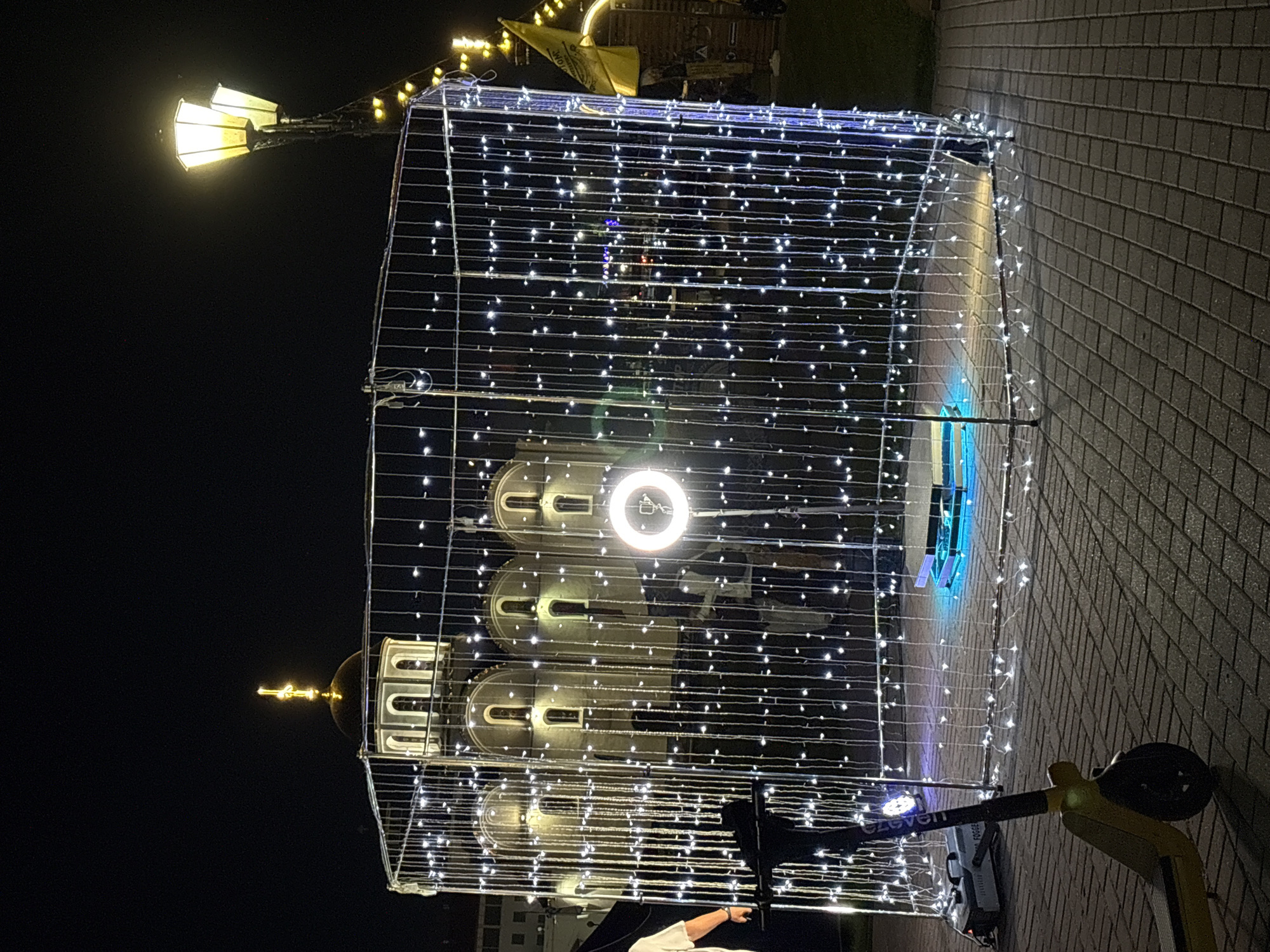 The image depicts an outdoor scene at night, featuring a large, illuminated structure made of metal framing and draped with numerous white string lights. This structure is rectangular and appears to be enclosed, with a circular ring light and a rotating platform visible inside. To the left of the structure, a yellow electric scooter is parked on a paved walkway. In the background, a building with a golden dome and arched windows is visible, suggesting a religious or historical landmark. A tall lamppost with two illuminated fixtures stands to the right of the frame, and a yellow flag is also present in the background. The overall impression is of a festive or event-related setup in a public urban space. Based on the provided context, this scene is located in the city of Vitsebsk, Belarus.
