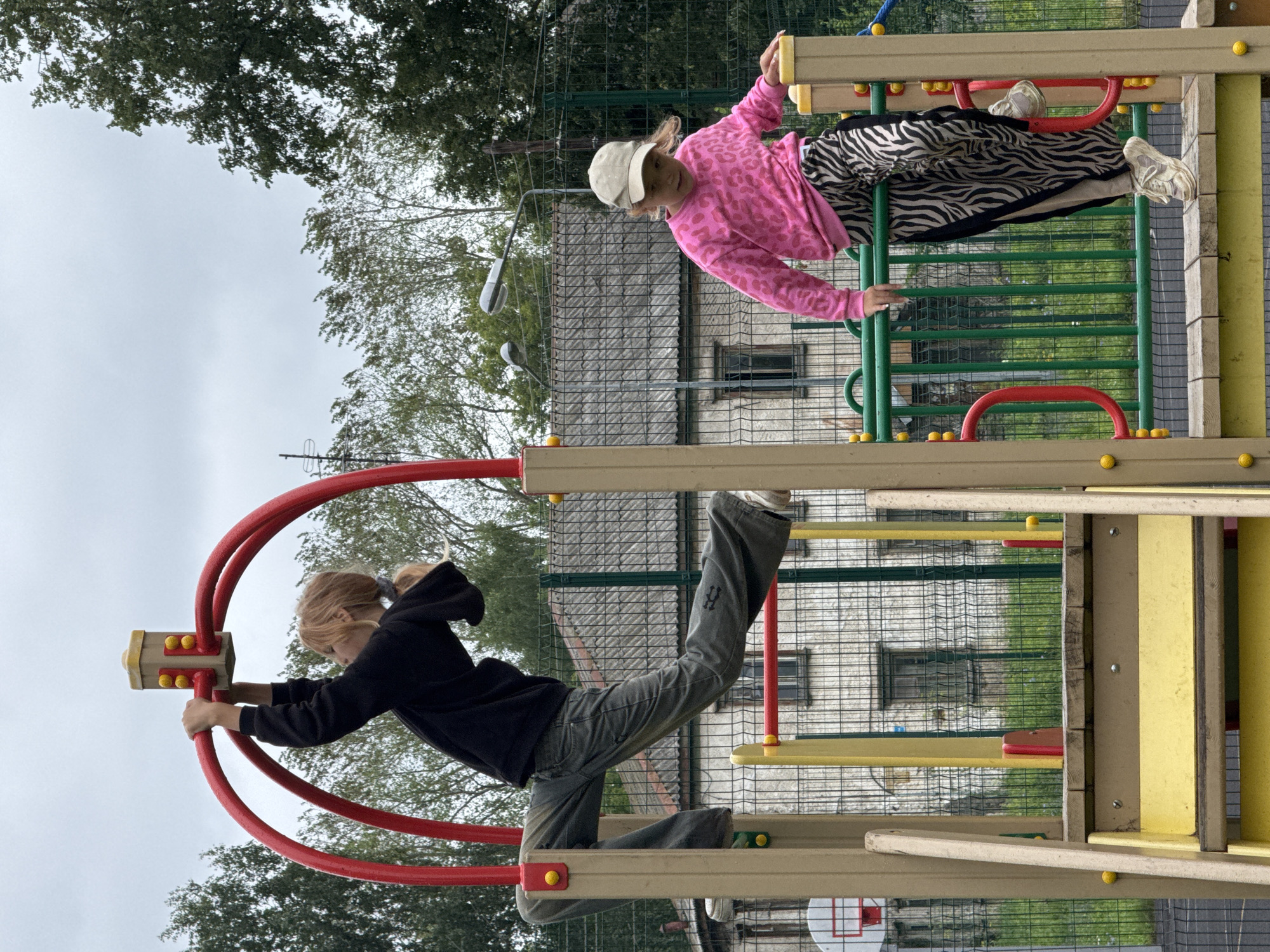 Two young girls are playing on a colorful playground structure. The girl on the left, with blonde hair and wearing a black hoodie and grey pants, is hanging from a red arch, suspended with one leg extended. The girl on the right, wearing a pink leopard-print sweatshirt, a beige cap, and black and white zebra-striped pants, is climbing on a wooden platform. The playground is situated in a fenced area with a basketball hoop visible to the left. In the background, there are trees and a two-story building with a grey, corrugated roof. The sky is overcast. This playground is located in the city of Chaikovsky, Russia.