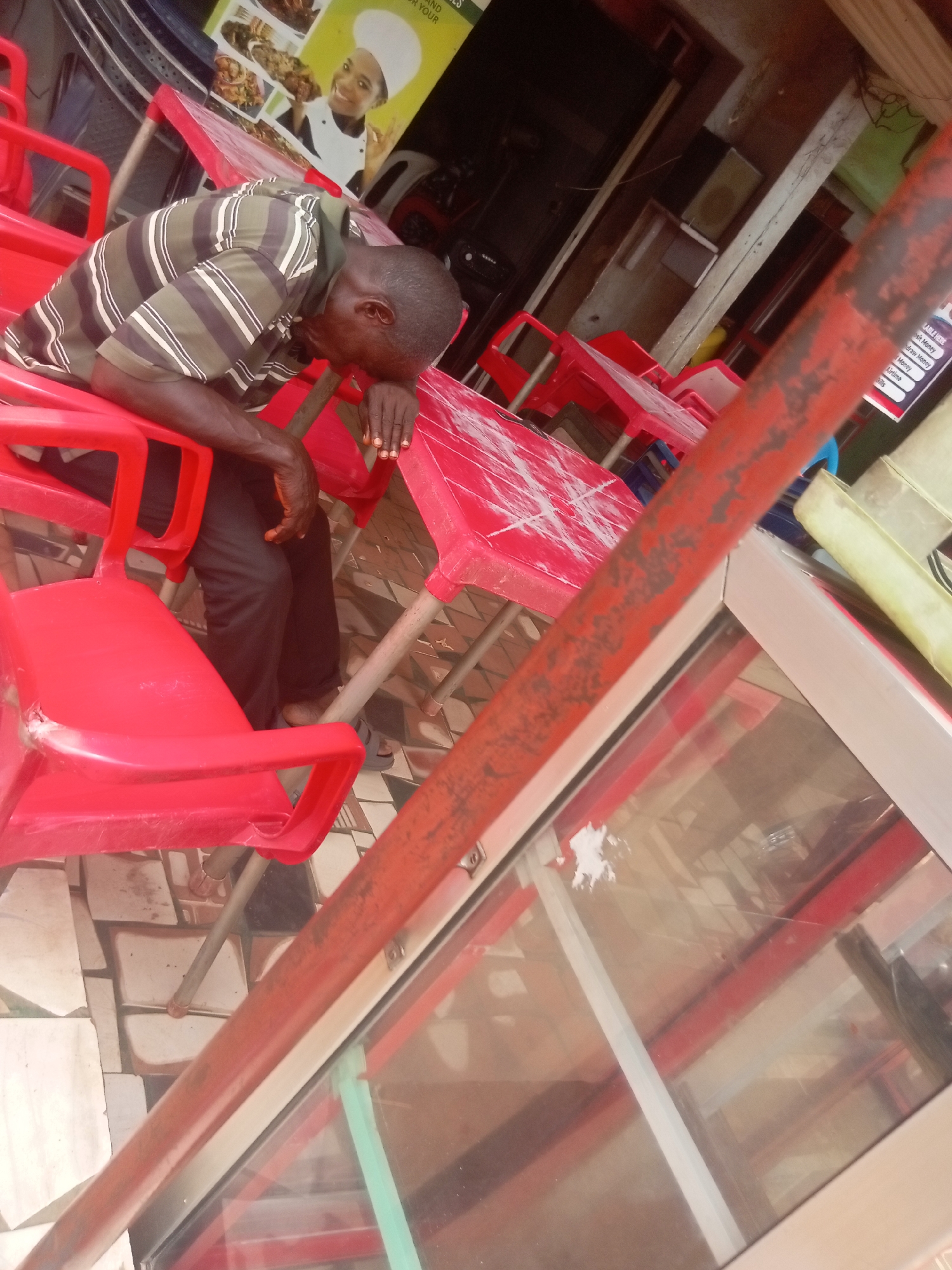 A man rests on a red plastic chair, his head on a red plastic table in Nkpor, Nigeria. He wears a striped shirt and black pants. The area appears to be an outdoor dining or seating area of a restaurant, with multiple red plastic chairs and tables. A menu board with a picture of a chef is visible in the background, and a metal framed glass window is in the foreground. The floor is tiled with brown and white patterns.