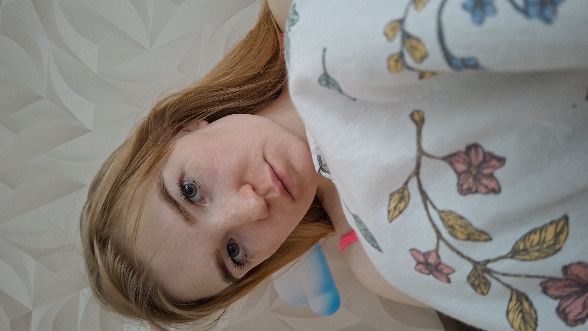 A low-angle portrait shows a fair-skinned woman with freckles. She has light-brown hair and light-blue eyes. Her expression is neutral. Part of a floral-patterned garment is visible on the right side of the image, a clear object with blue liquid can be seen just left of it. A textured white wall is in the background. The photo appears to have been taken indoors in Chaikovsky, Russia.