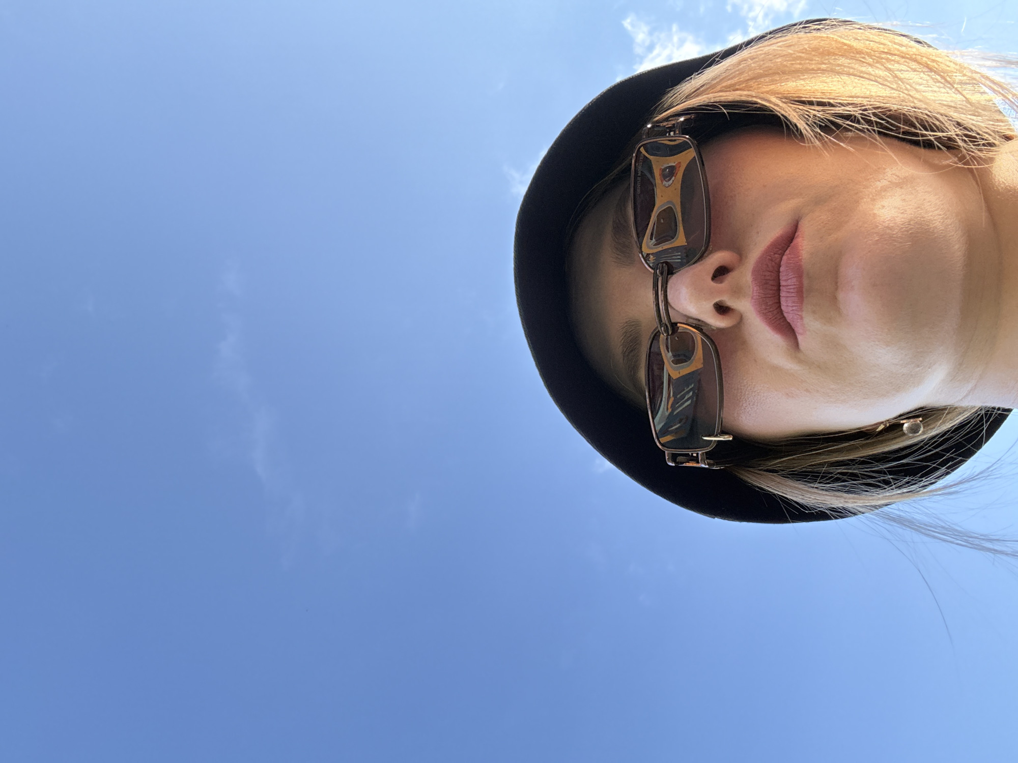 The image is a low-angle, close-up shot of a person wearing sunglasses and a dark bucket hat against a clear blue sky with some wispy clouds. The person's face is partially visible, with their lips slightly parted. The sunglasses reflect a yellow object, possibly a building or a sign. The hair is blonde and styled loosely. The setting appears to be outdoors during the day. No specific landmarks or indications of the city of Kazan, Russia are present in the image.