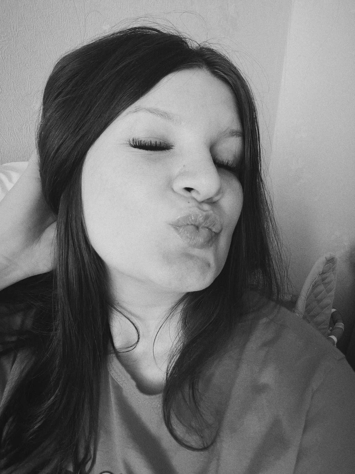 This black and white photograph, captured in Tobolsk, Russia, presents a close-up portrait of a young woman. Her long, dark hair frames her face, with strands falling over her shoulders and chest. She is depicted with her eyes closed and her lips puckered into a playful "kissing" expression, conveying a lighthearted and affectionate mood. One of her hands is visible, casually resting on the back of her head. She appears to be wearing a casual top or t-shirt. The background consists of a plain, textured wall, suggesting an indoor setting. Soft, diffused lighting illuminates the scene, creating gentle shadows. The image has a slightly grainy quality. There is no visible text.