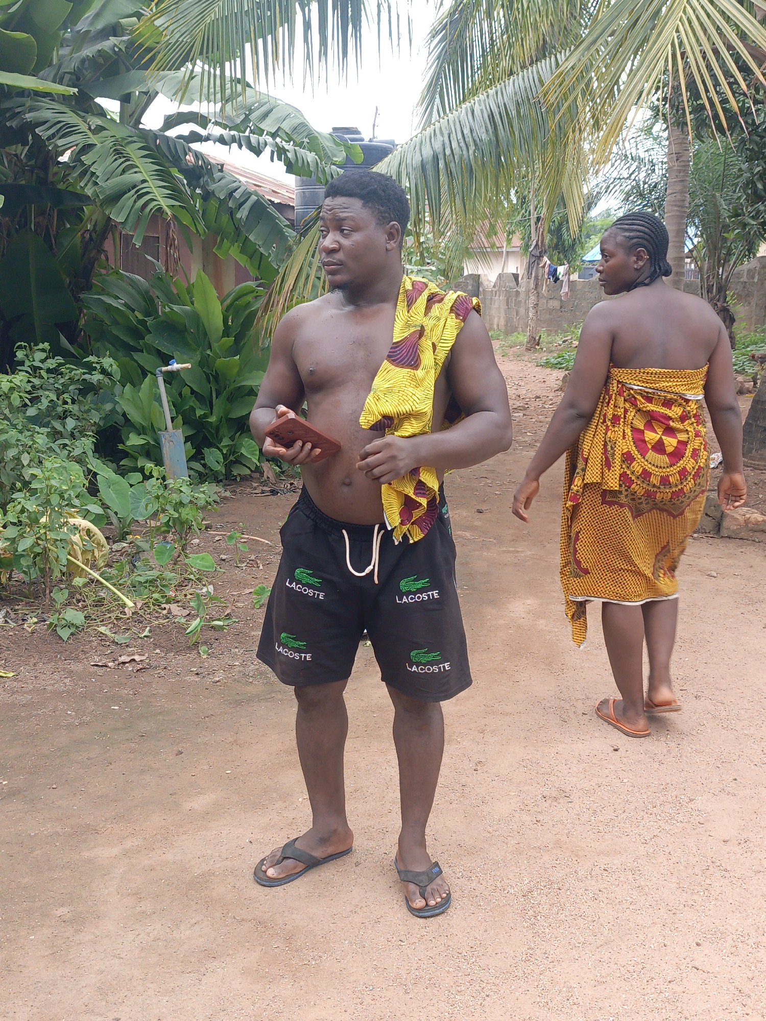 In Akwanga, Nigeria, a shirtless man stands on a dirt path, holding a reddish-brown smartphone in his right hand. He wears black shorts with two white "Lacoste" logos and is draped with a yellow and gold patterned fabric over his left shoulder. To his right, a woman walks away, also draped in a yellow and gold patterned fabric, secured around her chest and extending to her knees. Her hair is braided and pulled back. The environment is tropical, with lush green banana plants and palm trees in the background, and a simple fence made of grey bricks. The ground is a mix of dirt and small stones.