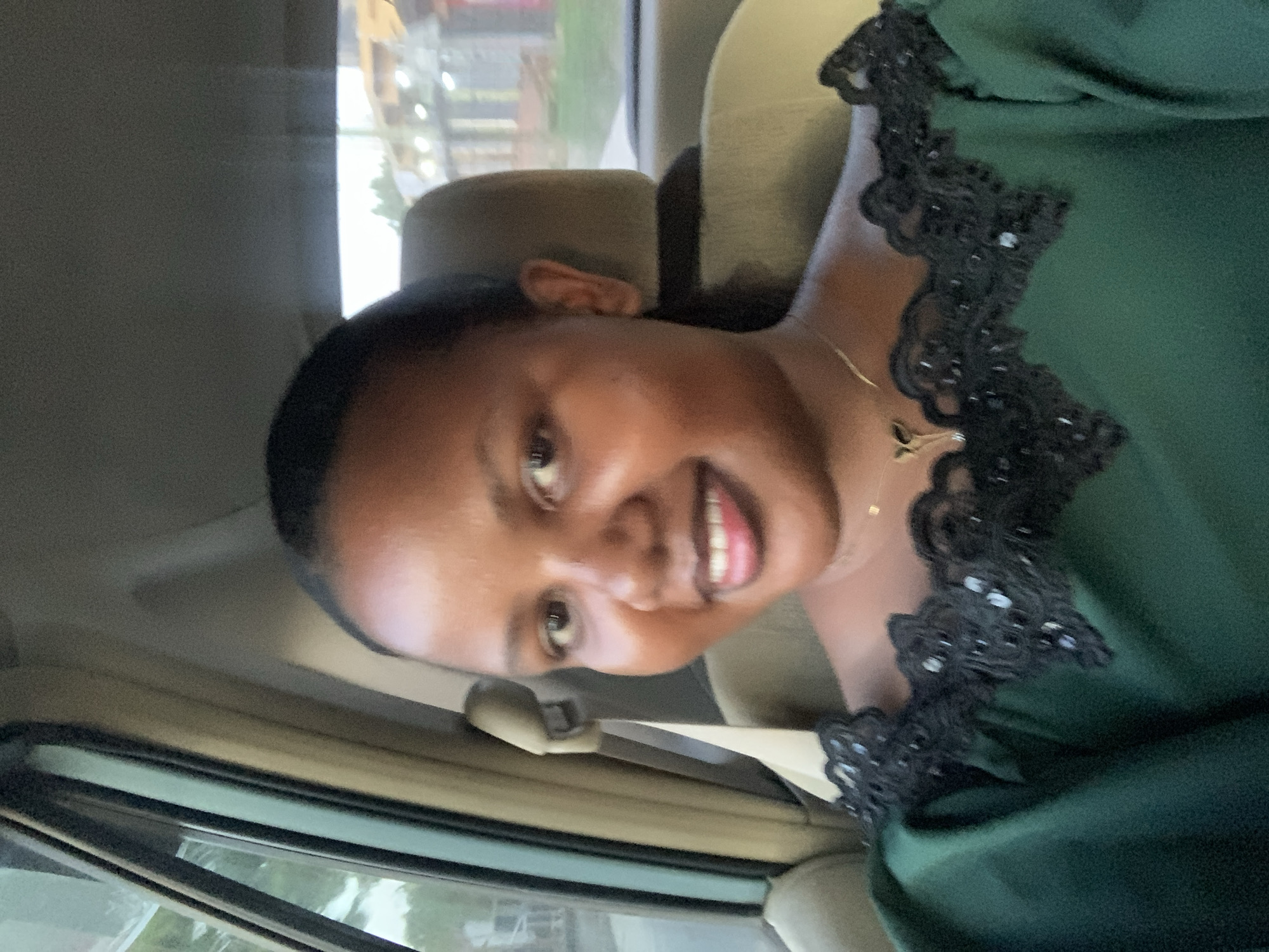 A woman is in the passenger seat of a car, smiling at the camera. She is wearing a dark green dress with black lace trim. A gold necklace is visible on her neck. The car appears to be moving, as indicated by the blurred scenery visible through the car window. The location of Dar es Salaam, Tanzania, cannot be ascertained from the photograph.