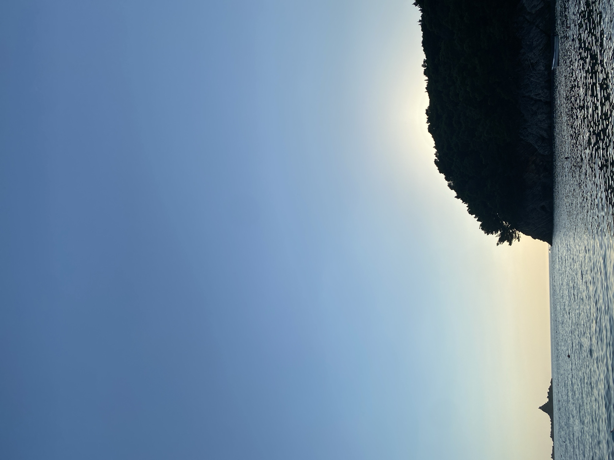 This is a low-angle, vertical shot of a serene seascape with a forested island and a cliffside in Petrovac centar, Montenegro. The sky is a gradient of clear blue transitioning to a pale yellow near the horizon. The water is calm with a ripple effect, reflecting the light. On the left, a small, rocky island with a structure on top is visible. In the center-right, a dark, forested cliffside dominates the frame, with the sun setting behind it, creating a silhouette. A small white boat is moored near the base of the cliff. A few buoys are scattered in the water. The overall impression is one of peaceful natural beauty.