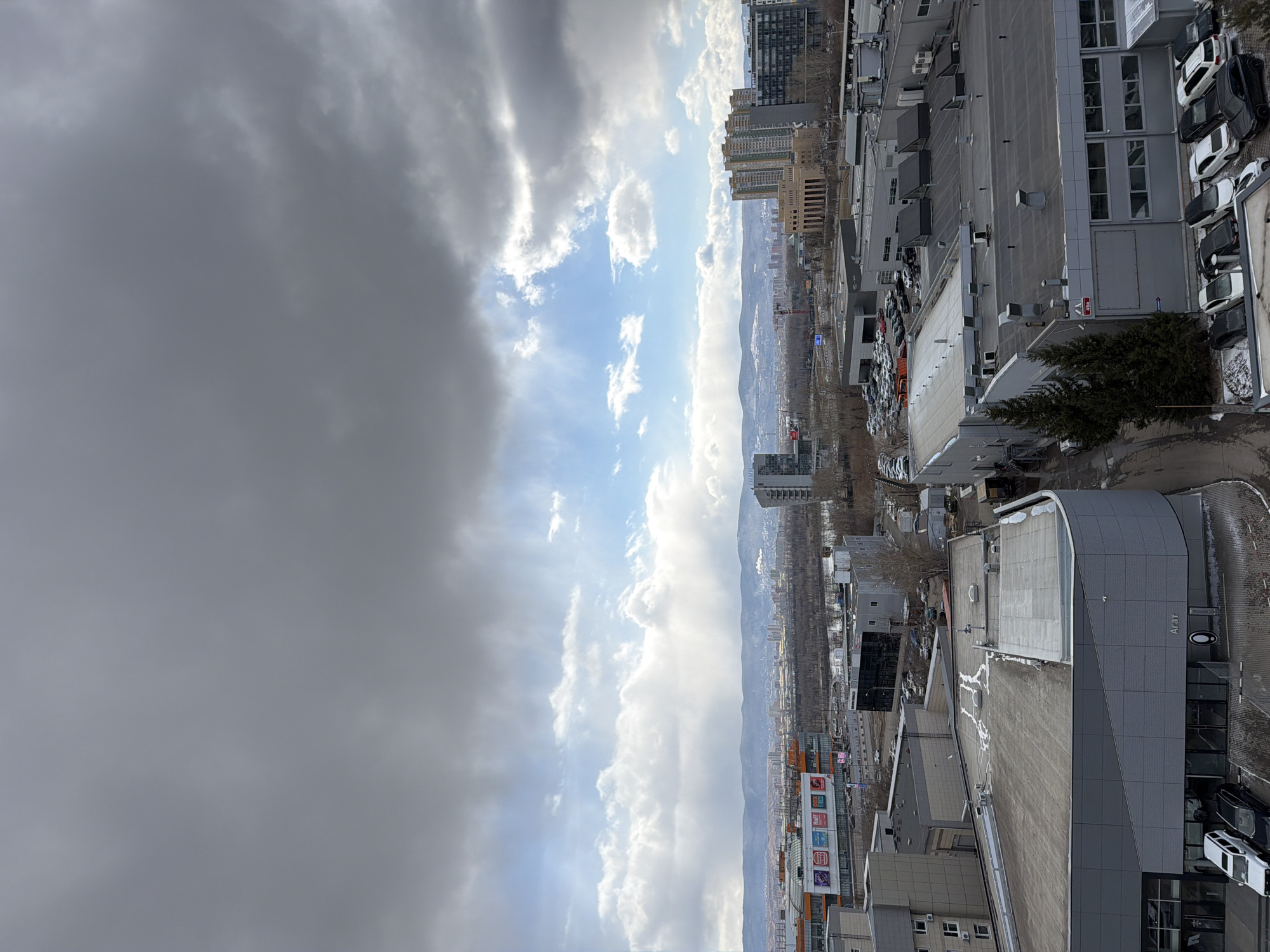 The image depicts a view from a high vantage point in Krasnoyarsk, Russia. The right side of the frame is dominated by buildings and structures with gray rooftops, parking areas, and visible vehicles. Snow is present on some surfaces. The left side presents a view of the sky, which is split between dark, ominous clouds and patches of blue sky with white clouds. In the distance, a city skyline is visible against the backdrop of rolling hills or mountains. The photo is taken during the day, and the overall scene suggests an urban environment on a day with mixed weather conditions.