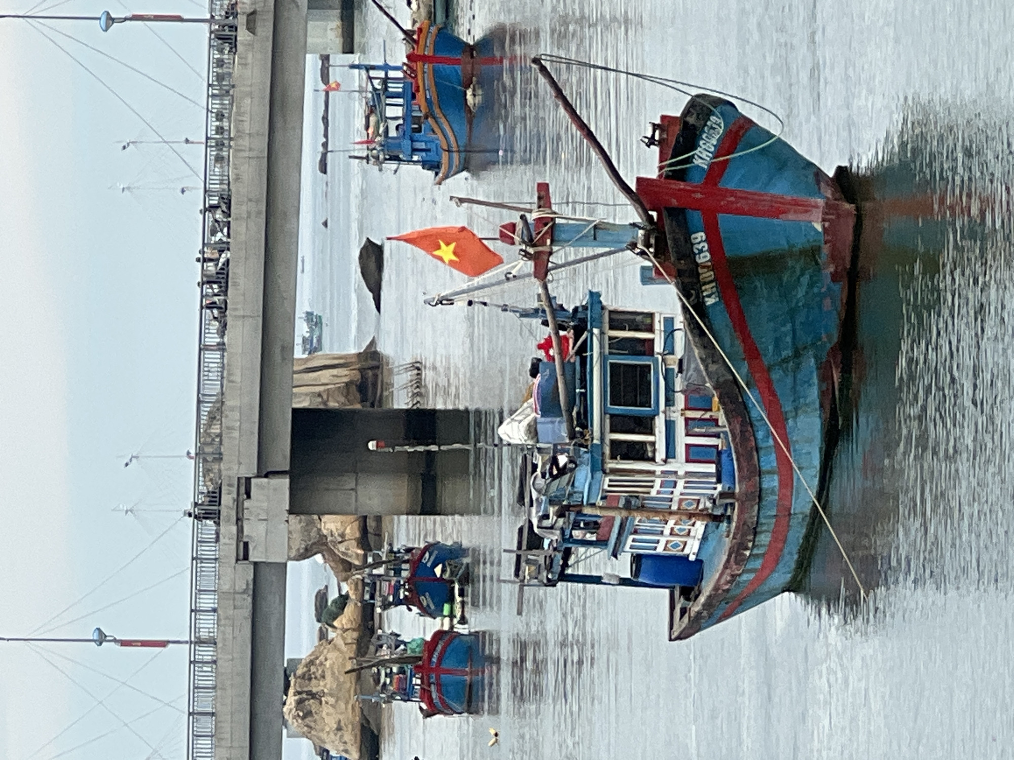 The image shows a wide shot of a harbor in Nha Trang, Vietnam, with several fishing boats docked in the foreground and mid-ground. A large concrete bridge spans the water in the background, with cars and pedestrians visible on its deck. The water is calm, with reflections of the boats and the sky. The sky is overcast, suggesting a cloudy day.
In the foreground, a blue and red fishing boat is prominently featured, its hull partially submerged in the water. The boat has a cabin with windows and a Vietnamese flag flying from a mast. The number "KH 09639" is visible on the bow of the boat.
In the mid-ground, two more fishing boats are visible, also painted in blue and red, with Vietnamese flags. Several rocks and a small island are scattered in the water around the boats. In the background, the bridge appears to be a significant structure, supported by large concrete pillars. A few people can be seen walking on the bridge.
The overall scene depicts a typical day in a Vietnamese fishing harbor, with a blend of traditional fishing vessels and modern infrastructure. The image captures the essence of maritime life in Nha Trang.